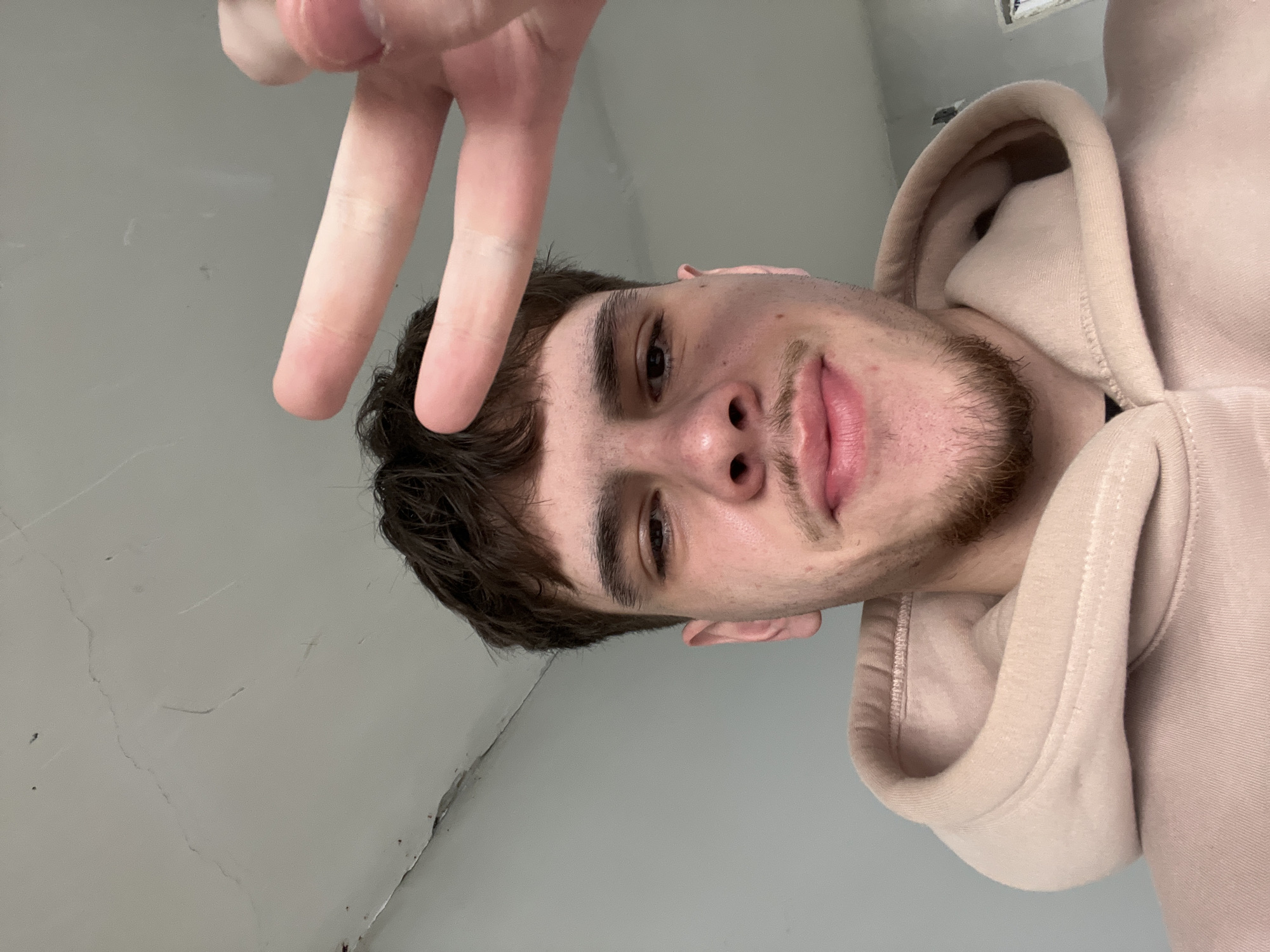 This close-up, upward-angled image features a young adult male subject indoors. The subject has short dark brown hair, dark eyes, and facial hair consisting of a mustache and a goatee. He is clad in a light beige hooded sweatshirt. His right hand is prominently displayed in the upper right quadrant, forming a "peace sign" with the index and middle fingers extended. The subject's expression includes slightly parted lips in a subtle smirk, with direct eye contact toward the camera. The background consists of a plain light-colored ceiling and an adjacent wall. The ceiling exhibits visible cracks, minor peeling paint or plaster, and dark discoloration along the seam where it meets the wall in the upper left portion of the image. No definitive visual cues are present to establish a specific location within the city of Sofia, Bulgaria.