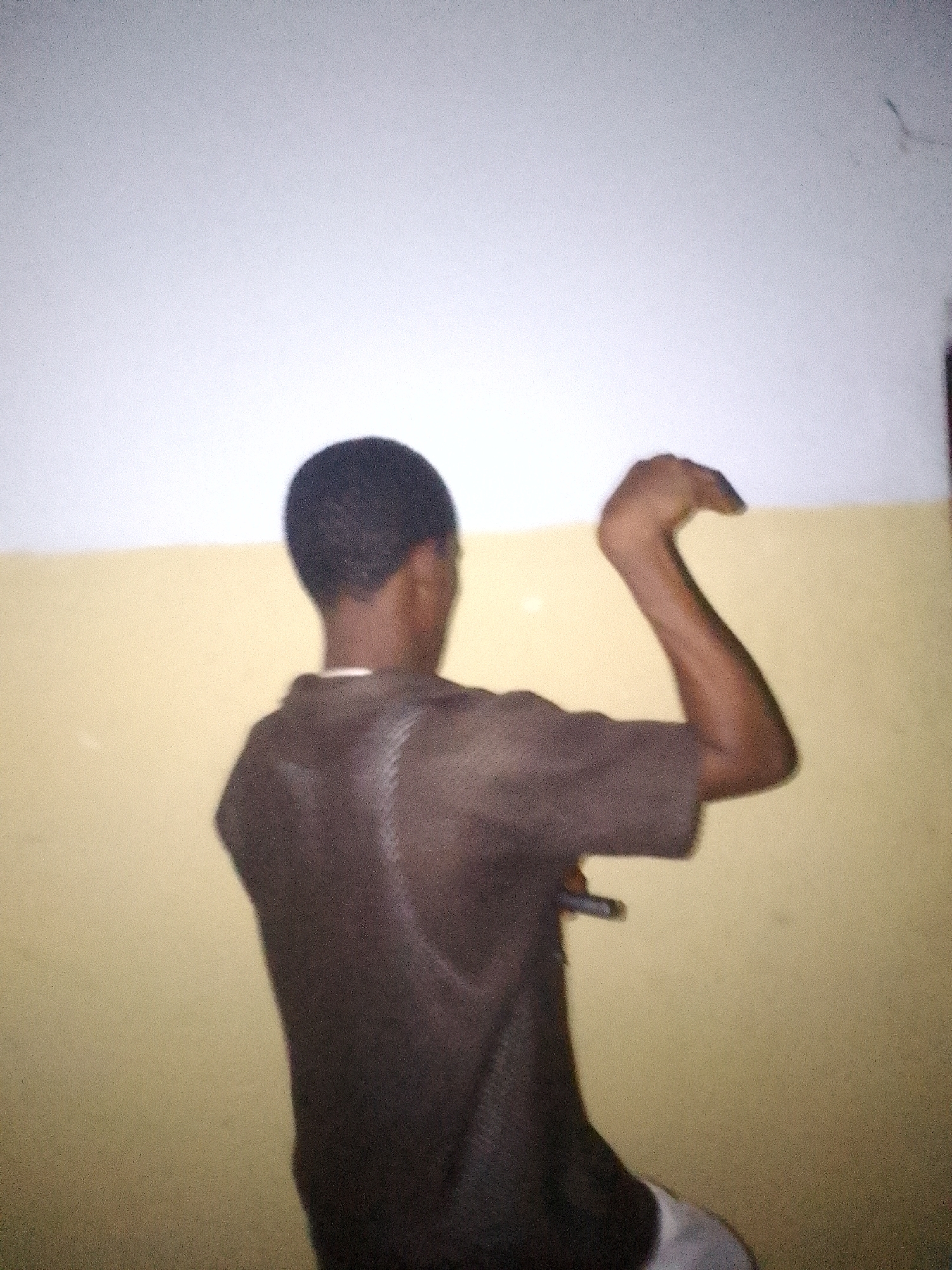 A photograph shows a person from the back, wearing a dark brown t-shirt, standing in a room with a wall that is two-toned, with a white upper section and a yellow lower section. The person has short, dark hair, and their right arm is raised, bent at the elbow. The location is unknown.