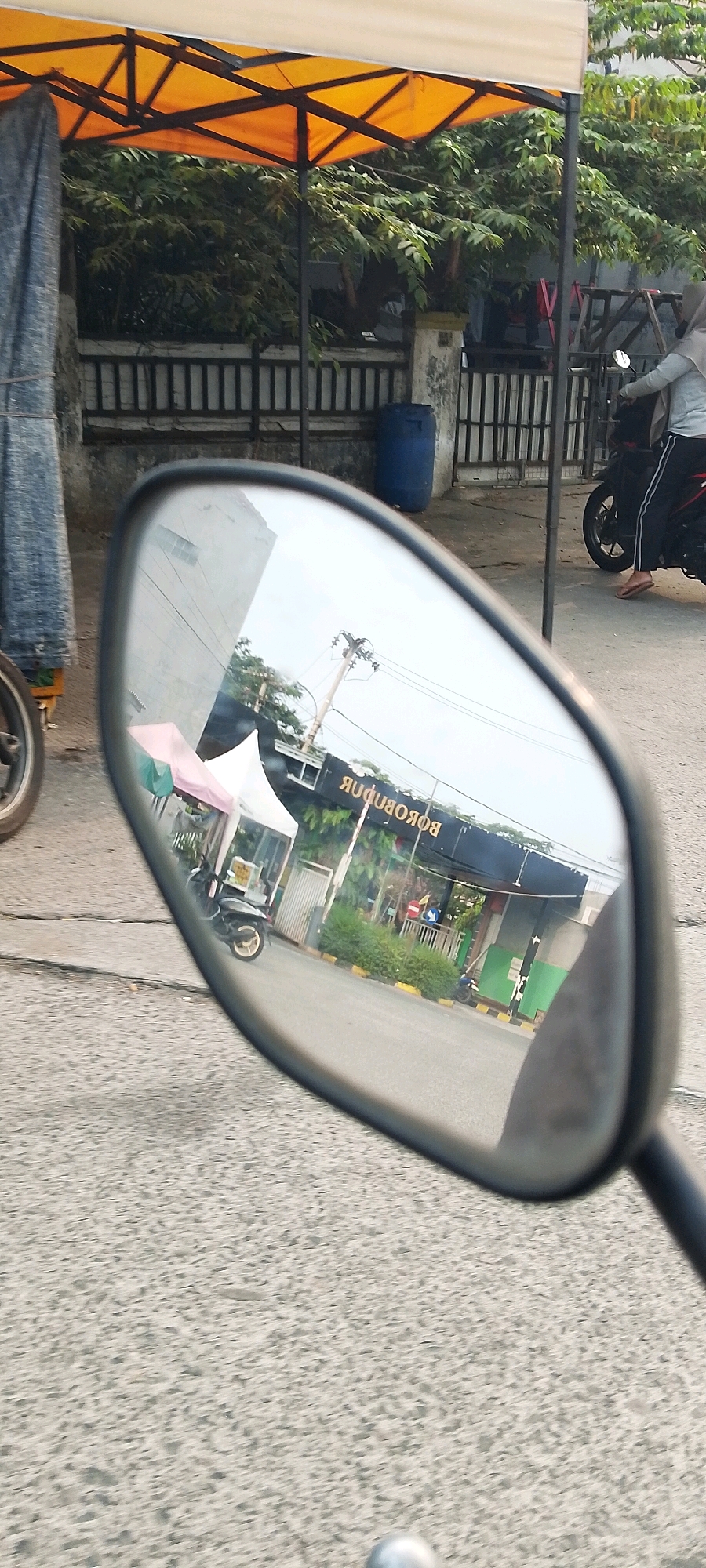 The image shows a reflection in a motorcycle's side-view mirror. The reflection captures a street scene with a building featuring a sign that reads "BOKOBOBOK" in reverse. There are two white tents with pink and green coverings, possibly stalls, and a parked motorcycle in front of the building. A power pole with wires is also visible. In the foreground of the actual image, the asphalt road and the motorcycle's frame are visible. In the background, an orange canopy tent and foliage can be seen, along with a blue barrel and a black metal fence. A person on a motorcycle wearing a hijab and a light-colored shirt is riding away from the camera. The scene appears to be outdoors in an urban environment in Satriamekar, Indonesia.