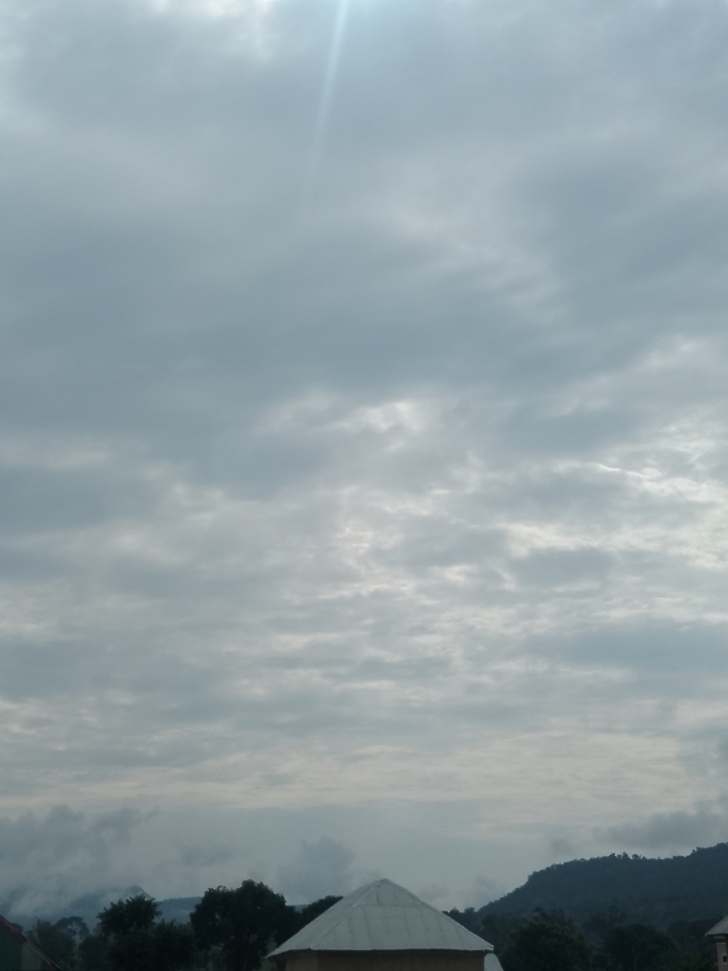 The image shows a cloudy sky over a rural landscape. In the foreground, the roof of a building is visible. Behind the building, there are trees and hills. The sky is filled with a thick layer of gray clouds, with a hint of sunlight breaking through at the top left. The overall scene is serene and appears to be an overcast day in a rural area, potentially in Serti, Nigeria, given the context.