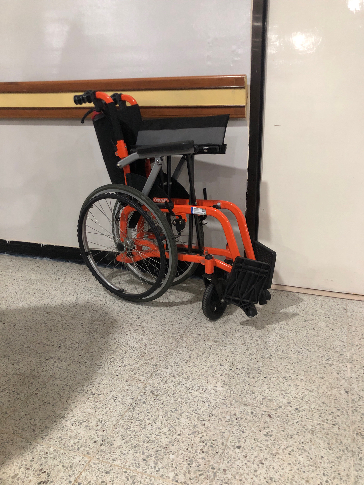 A folded orange wheelchair is leaning against a wall, positioned on a terrazzo floor. The wheelchair is predominantly orange with black accents, featuring large spoked wheels, a seat, and footrests. A wooden handrail runs horizontally along the wall behind the wheelchair. A dark vertical line, possibly a doorframe, is visible to the right of the wheelchair. The lighting suggests an indoor environment, casting shadows on the floor. The scene is devoid of people and any discernable indications of the city of Surabaya, Indonesia.