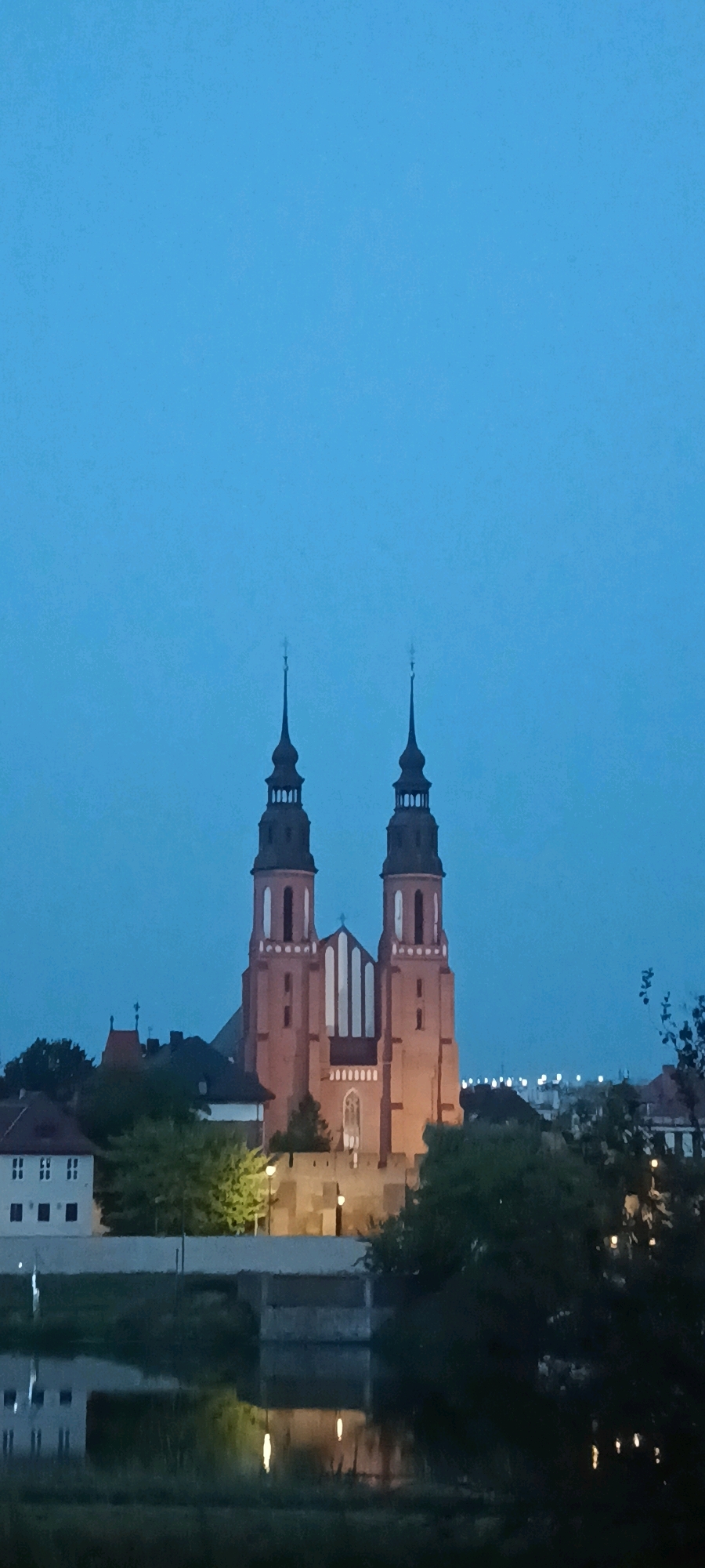 The image depicts the Opole Cathedral, also known as the Cathedral of the Holy Cross, situated in Opole, Poland. The architectural style is Gothic Revival, characterized by its twin spires and red brick facade. The cathedral is illuminated, suggesting it is either dawn or dusk, with a clear blue sky overhead. In the foreground, a body of water, likely the Oder River, reflects the lights of the city and the cathedral itself. The surrounding area features other buildings and trees, contributing to the urban landscape. The scene is peaceful and serene, highlighting the grandeur of the cathedral against the twilight sky.