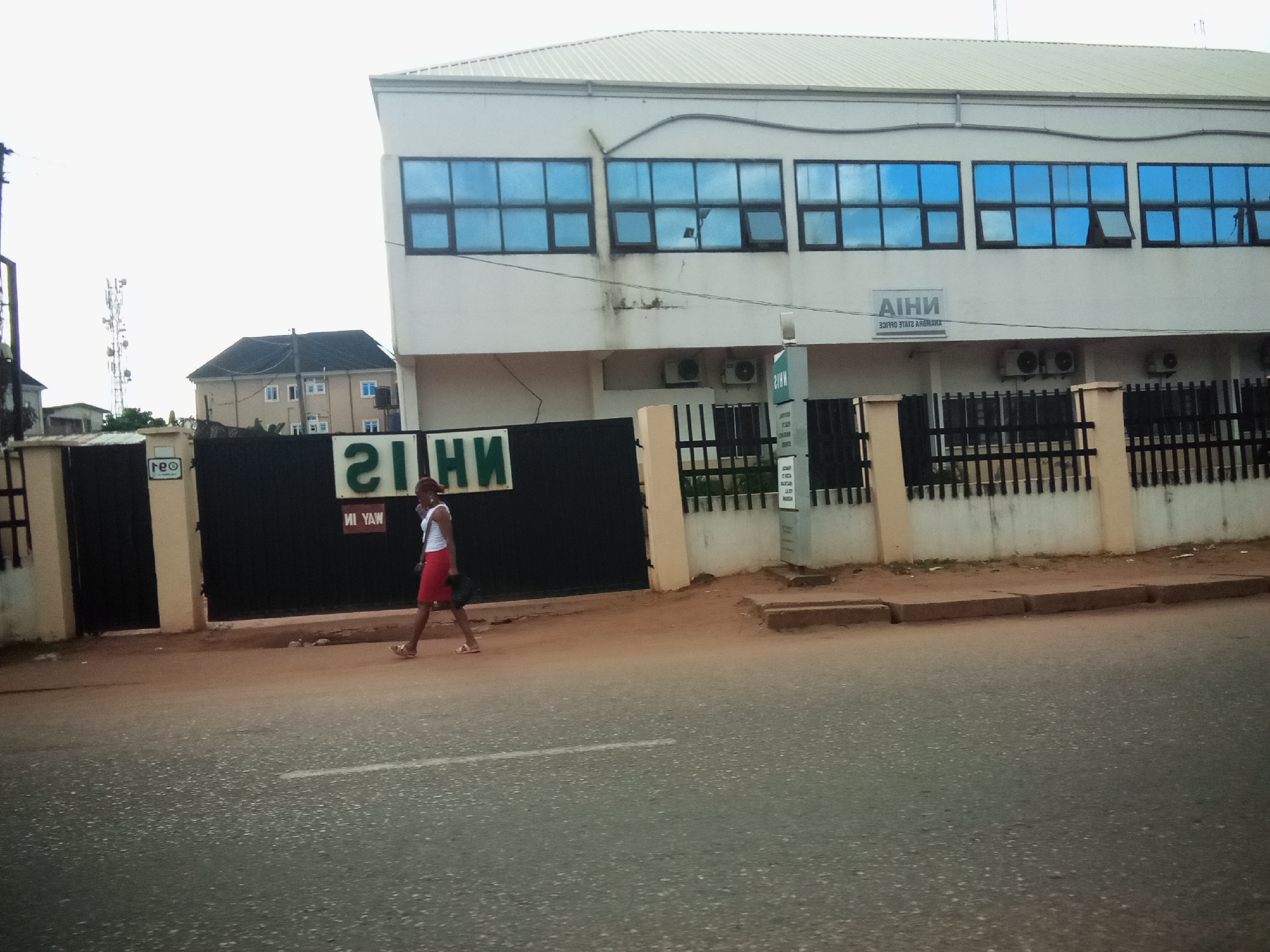 A medium-shot exterior view captures a street scene in Nibo, Nigeria, under an overcast sky. In the foreground, a paved road with a white dashed line runs horizontally across the frame. On the right side of the road, a woman wearing a white top and a red skirt walks from left to right, carrying a black bag. She has on light-colored sandals.
Behind the woman is a black metal gate with a prominent green and white sign displaying the text "21NH" in large lettering, followed by "NI YAW" in smaller lettering. To the right of the gate, a cream-colored building with a white roof stands. This building has multiple blue-tinted windows on its upper floor. A smaller sign above the windows reads "AIHN" with smaller text below that is difficult to decipher but appears to be an address or name. Two air conditioning units are mounted on the lower part of this building's facade. Beyond this building and to the left, a beige residential building with multiple windows and a dark roof is visible. A tall telecommunications tower is also seen in the distance. The ground area between the road and the buildings is a mix of sand and dirt, with low concrete curbs.