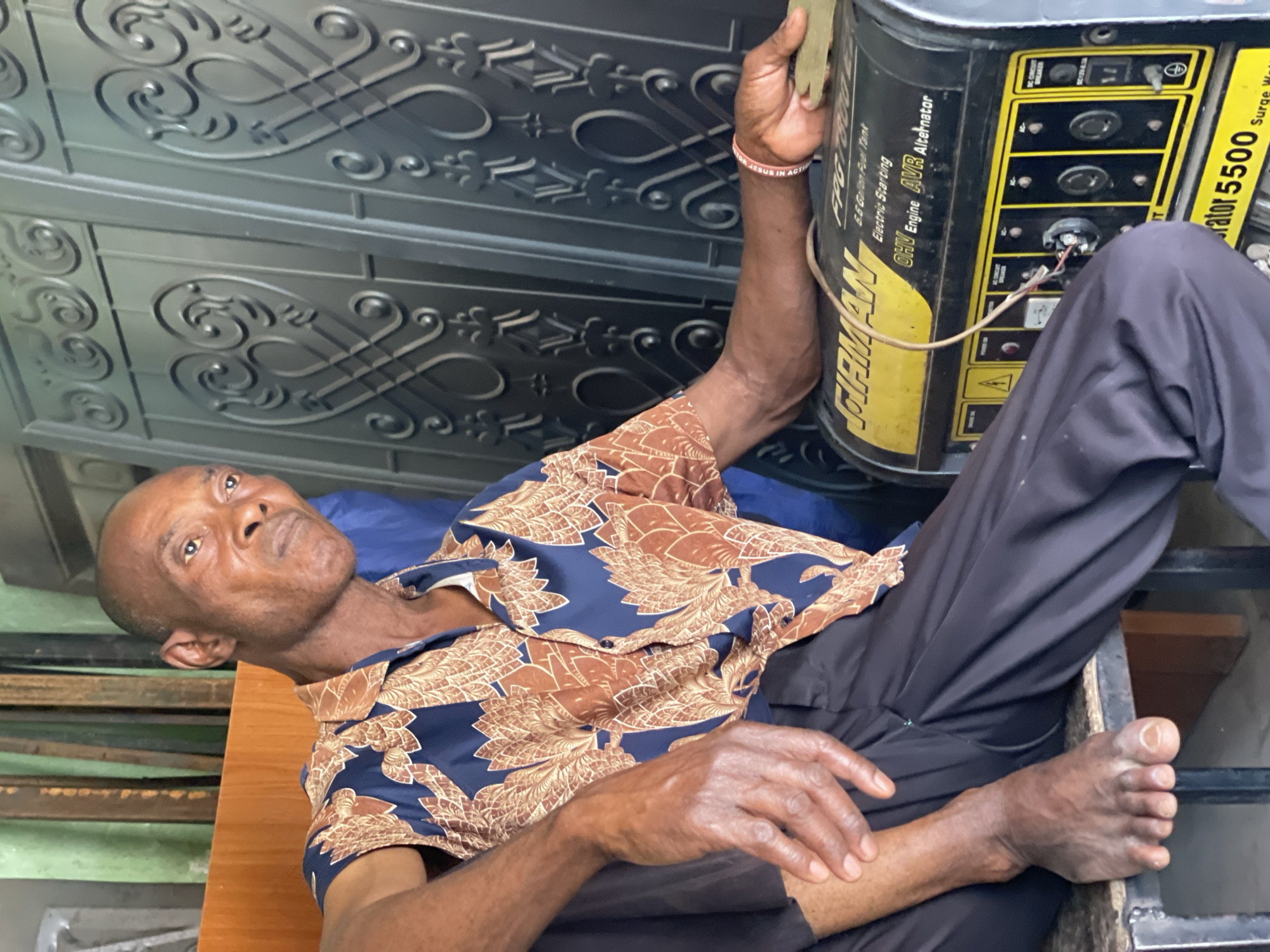 An elderly or middle-aged man with dark skin and a bald head is depicted resting or reclining in Zaria, Nigeria. He wears a short-sleeved blue shirt with intricate brown and gold patterns and dark trousers, and is barefoot. His gaze is directed upwards, conveying a calm or pensive expression.
He is positioned next to a prominent black and yellow portable generator. His right hand is near or resting on the generator, and on his right wrist, he wears a light-colored wristband bearing the text "JESUS IN ACTION." Visible text on the generator includes "FIRMAN", "FPG 7000E", "6.6 Gallon Fuel Tank", "Electric Starting", "OHV Engine AVR Alternator", "DC CIRCUIT BREAKER", "Generator 5500", and "Surge Watts". A wire is seen connecting to the generator's control panel.
The background features an ornate, dark metal gate or decorative wall with intricate scrolled patterns. To the man's left, a wooden surface, possibly a table or bench, is visible, and a blue cushion or fabric supports his head. The indirect lighting suggests an indoor setting or a shaded outdoor area. The scene captures a quiet moment of repose, with the generator indicating a practical aspect of daily life.