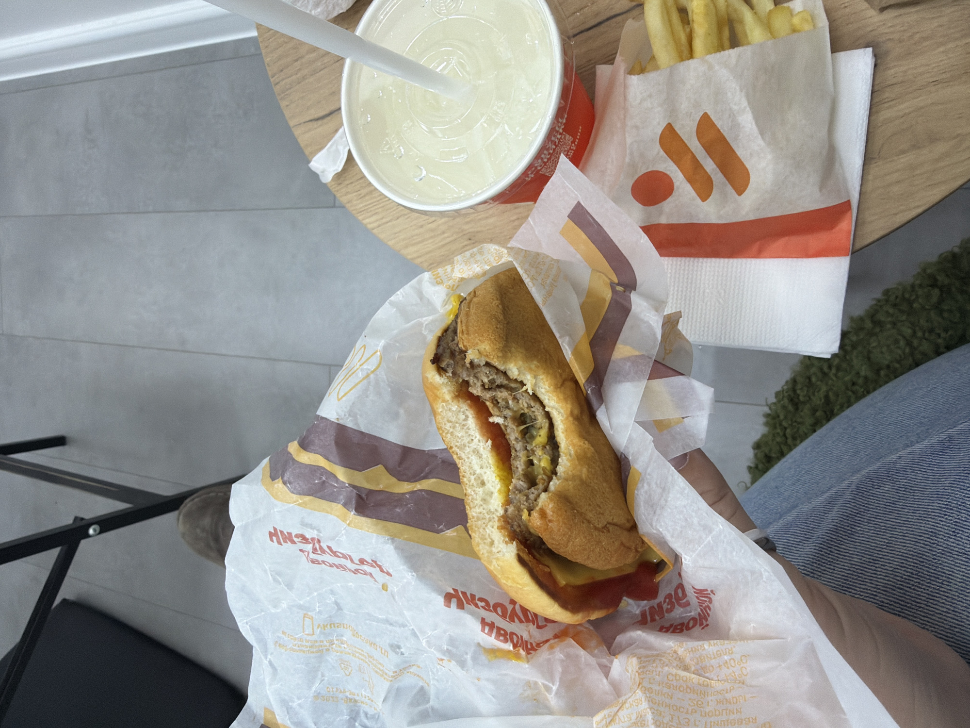 An overhead, first-person shot captures a fast-food meal set on a light wooden table, likely inside a restaurant or casual dining area. A person's hand, partially visible on the right, holds a half-eaten long burger or sandwich, which is wrapped in white paper featuring yellow and brown wavy stripes. The burger's interior reveals a meat patty, melted cheese, and a reddish sauce, possibly ketchup, on a sub-style bun.
To the left of the burger, a white paper cup with a red base contains a clear, light-colored beverage with ice and a white straw. To the right, a white paper bag filled with french fries rests on the table. The fries bag features a distinct orange logo: two vertical orange lines accompanied by a single orange dot to their left, above a horizontal orange stripe. This branding is characteristic of "Vkusno i Tochka," a Russian fast-food chain.
The wrapper of the burger and other items display Cyrillic text, including "ВКУСНО — И ТОЧКА" (Vkusno i Tochka), and partially visible "Чизбургер Де Люкс" (Cheeseburger De Luxe), "АКЦИЯ" (Action/Promotion), and "ПОДАРОК" (Gift).
The background shows a grey tiled floor, a white baseboard, and dark metal furniture, possibly a chair leg or table frame. Part of a person's leg clad in blue denim jeans is visible on the far right, next to a fuzzy green object, possibly a rug or part of a seat. The scene depicts a casual dining experience, with the individual consuming their meal. The branding confirms the context of Moscow, Russia.