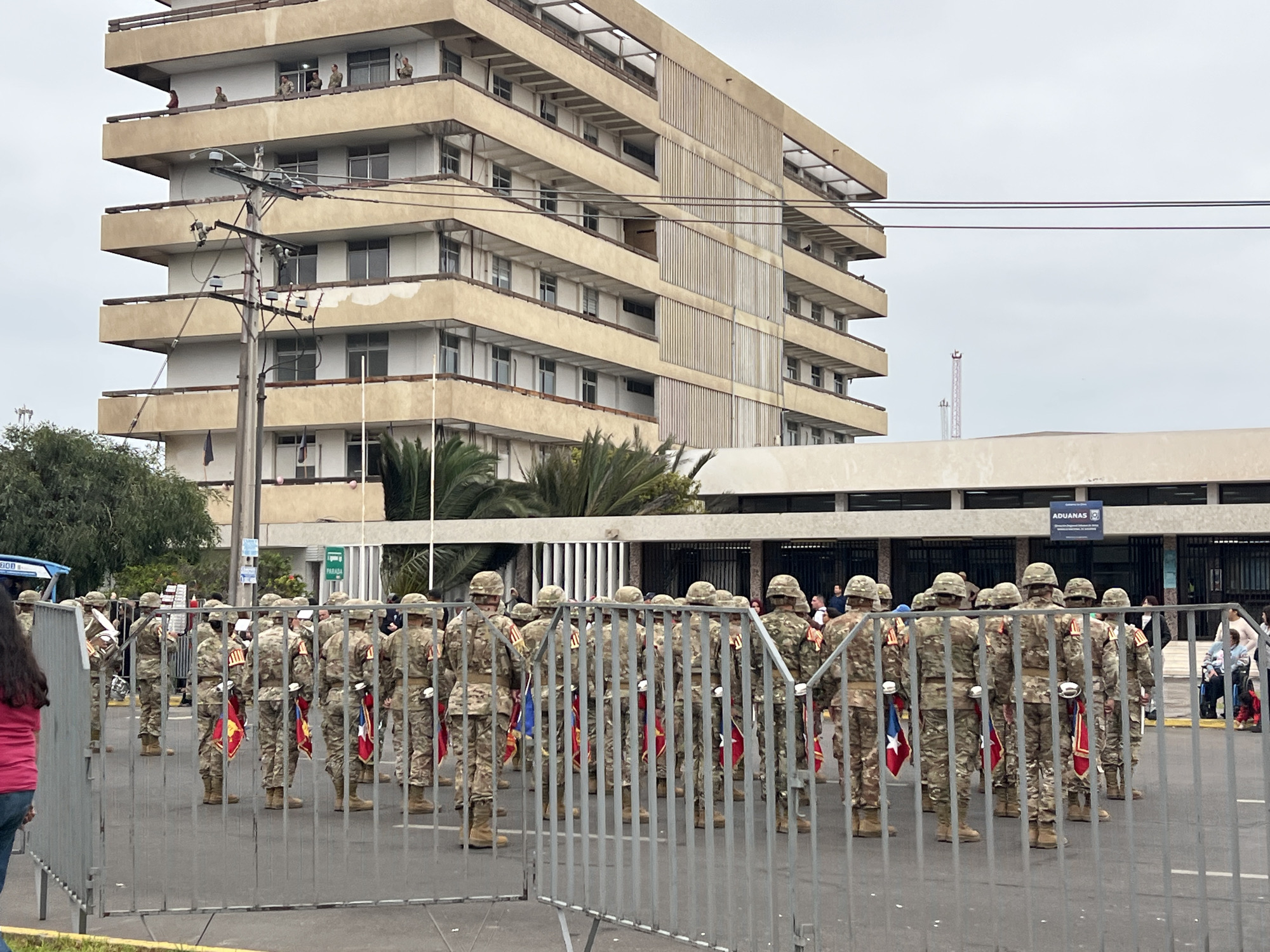 The image depicts a military parade or ceremony taking place in front of the Aduanas building in Arica, Chile. A formation of soldiers in camouflage uniforms and helmets stands in formation, facing away from the camera, holding flags and musical instruments. They are separated from the viewer by a metal fence. Behind the soldiers, the Aduanas building is visible, a large, multi-story structure with balconies and windows. A power pole and electrical wires are also prominent in the foreground. A few civilians can be seen in the background, including some on balconies of the building and a woman in a pink shirt in the lower left corner. The sky is overcast.