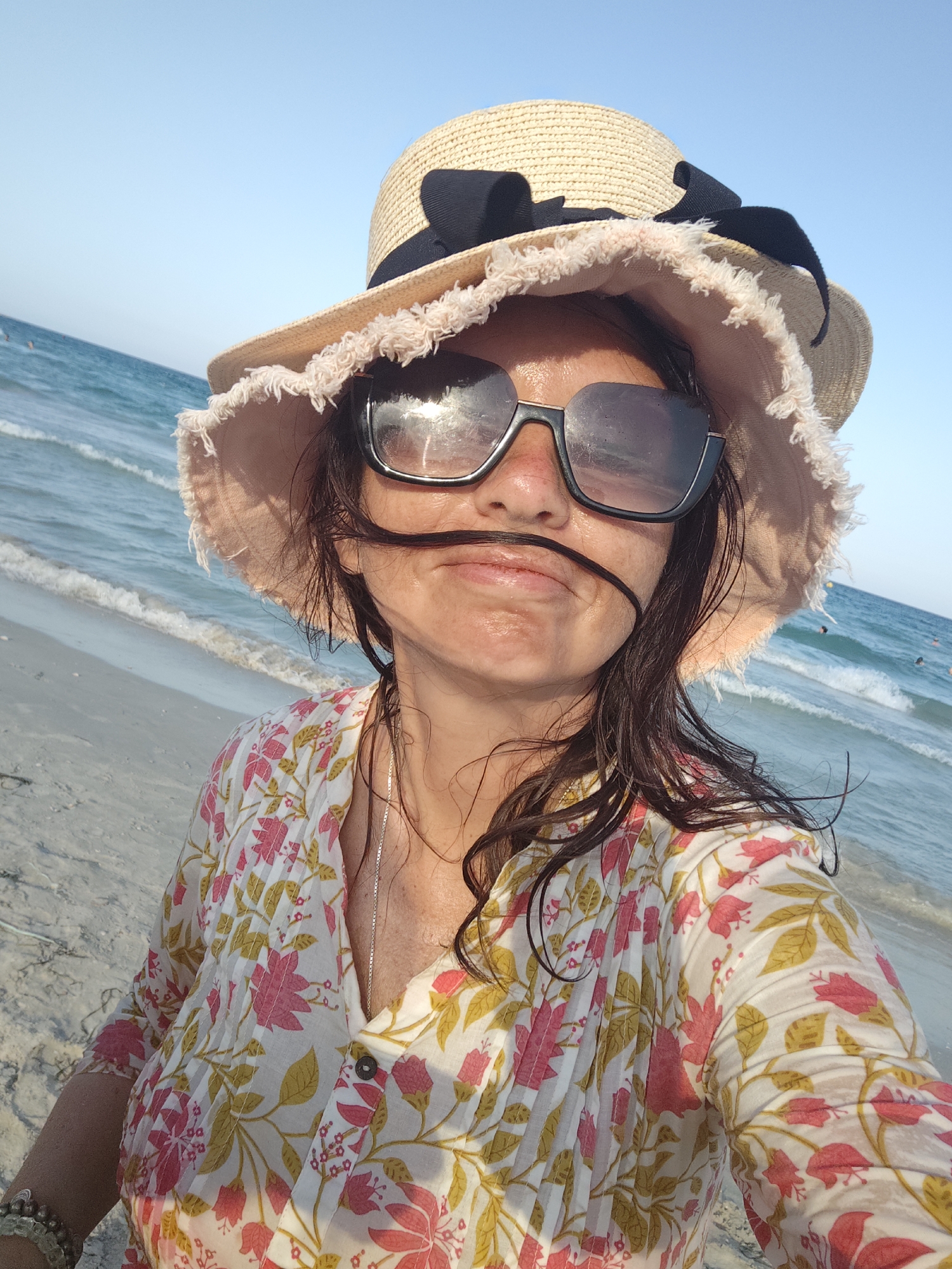 A woman is taking a selfie on a beach in Mahdia, Tunisia. She is wearing a wide-brimmed straw hat with a black ribbon, large sunglasses, and a white blouse with a floral print. Her hair is dark and wet, and a strand is blowing across her face. The ocean is visible in the background, with gentle waves lapping at the shore. The sky is clear and blue.