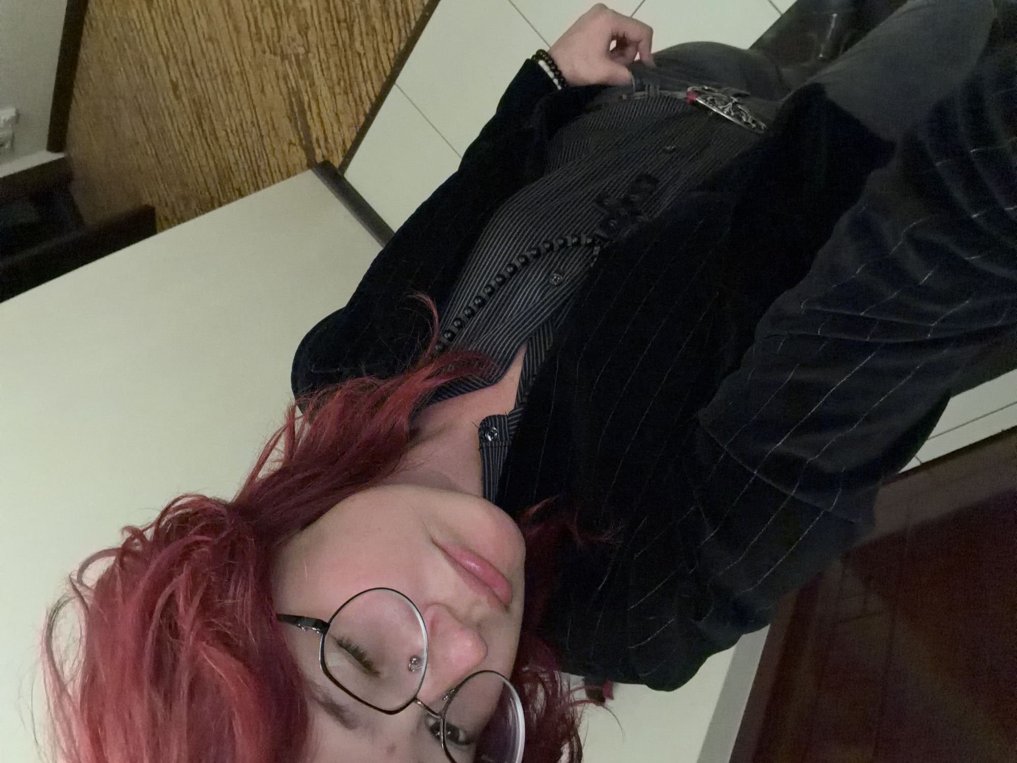 A person with pink hair and glasses is posing for a selfie. They are wearing a black pinstripe jacket over a pinstripe shirt and grey pants. The background appears to be an indoor setting with a light-colored wall and a wooden textured surface. There is no indication of the city of Chelyabinsk, Russia, or any specific urban environment.