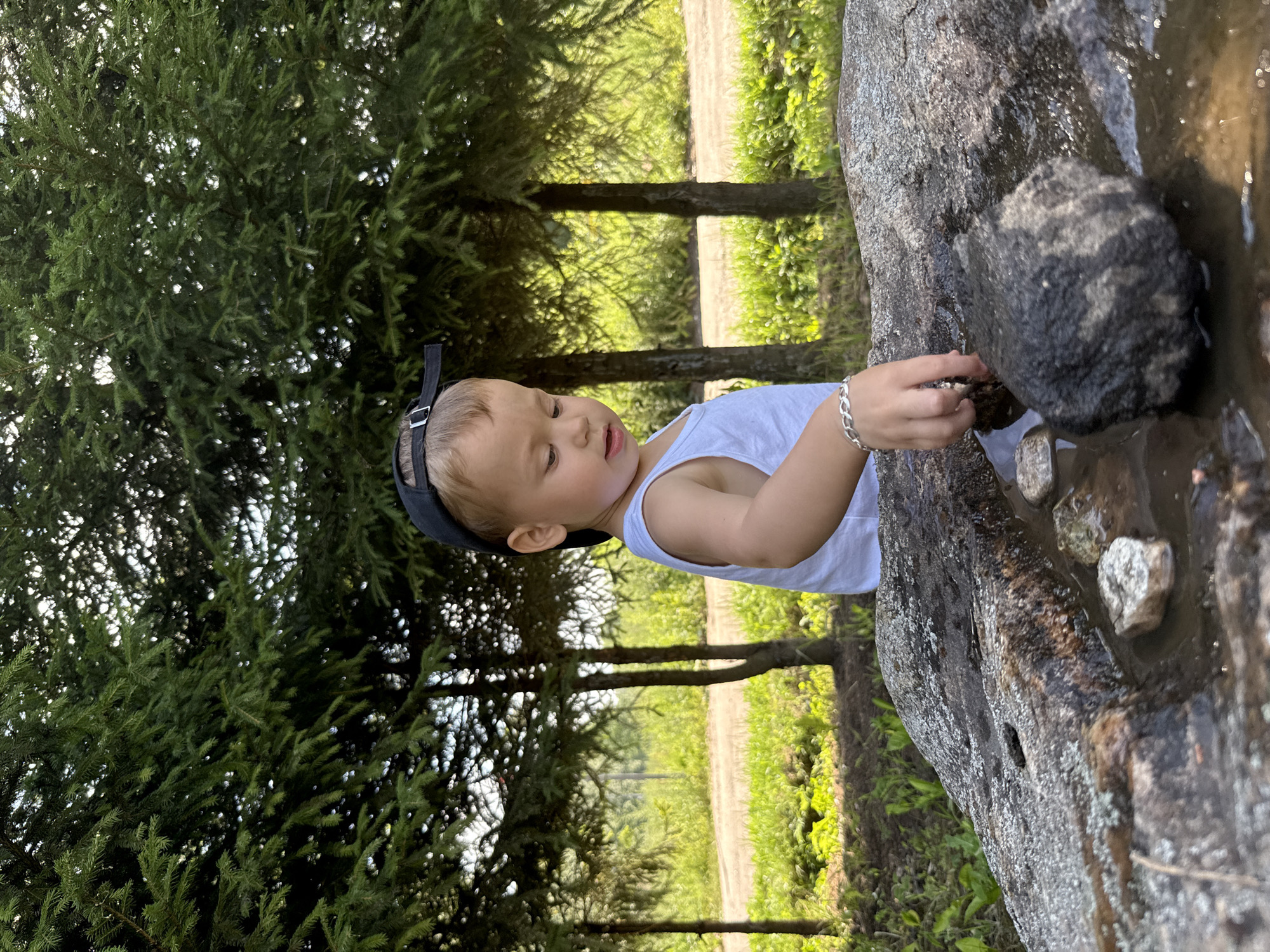 In Rusanovo, Russia, a young boy wearing a white tank top and a black baseball cap is playing with stones in a puddle on a large rock. He is reaching into the water with his right hand, which is adorned with a silver bracelet. The rock is rough and textured, with patches of moss and lichen. Behind him, a line of evergreen trees forms a dense backdrop. Sunlight filters through the branches, creating dappled light on the forest floor. The ground is covered with green grass and foliage. A dirt road is visible in the distance.