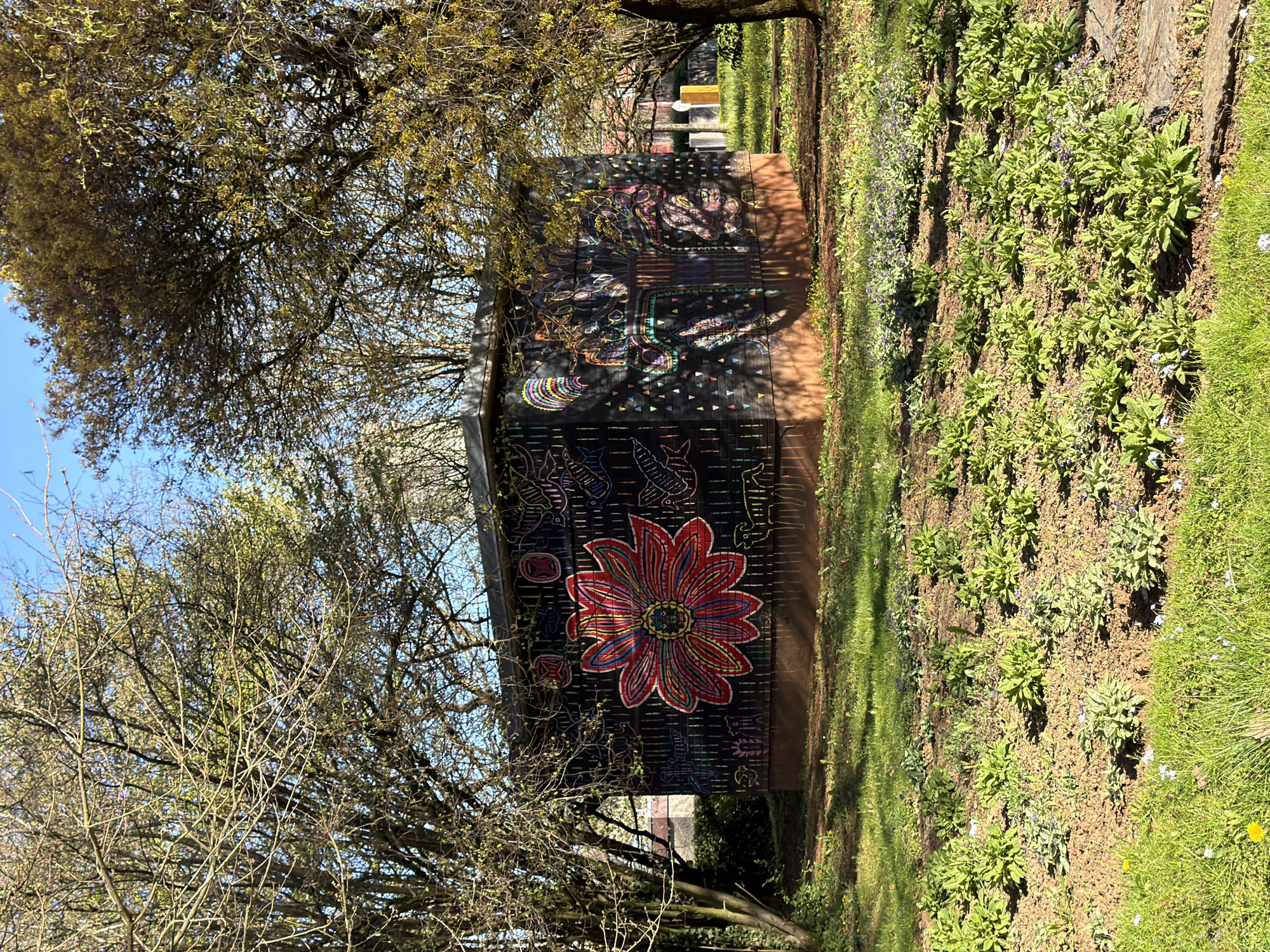 The image presents a view of a shed or small structure with a mural painted on its side. The structure is located in a park or garden-like setting in the city of Esch-sur-Alzette, Luxembourg. The mural features a variety of colorful designs, including a large red flower, along with other abstract and geometric shapes. The shed is bordered by a flower bed or garden area with various plants and greenery. Overhanging trees with sparse foliage frame the structure. The environment appears to be outdoors on a sunny day.