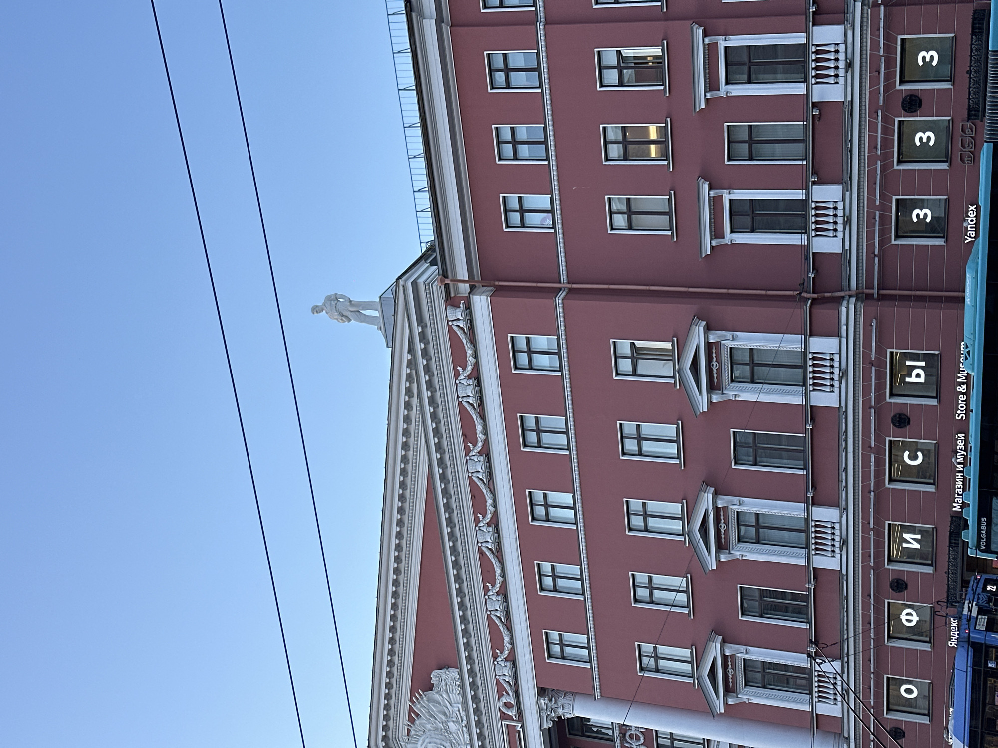 The image shows a building in Saint Petersburg, Russia. It is a multi-story building with a red facade and white architectural details. There are numerous windows on each floor, some with decorative molding. On top of the building is a statue. The building is identified as a store and museum with the word "Магазин и музей" written in Russian, and "Store & Museum" in English. A Volgabus number 22 is visible on the right side. Signage with numbers "3", "3", "3" and "Ы" is also present. The word "Yandex" can be seen on the building too. Power lines are visible in the sky. The sky is clear and blue.