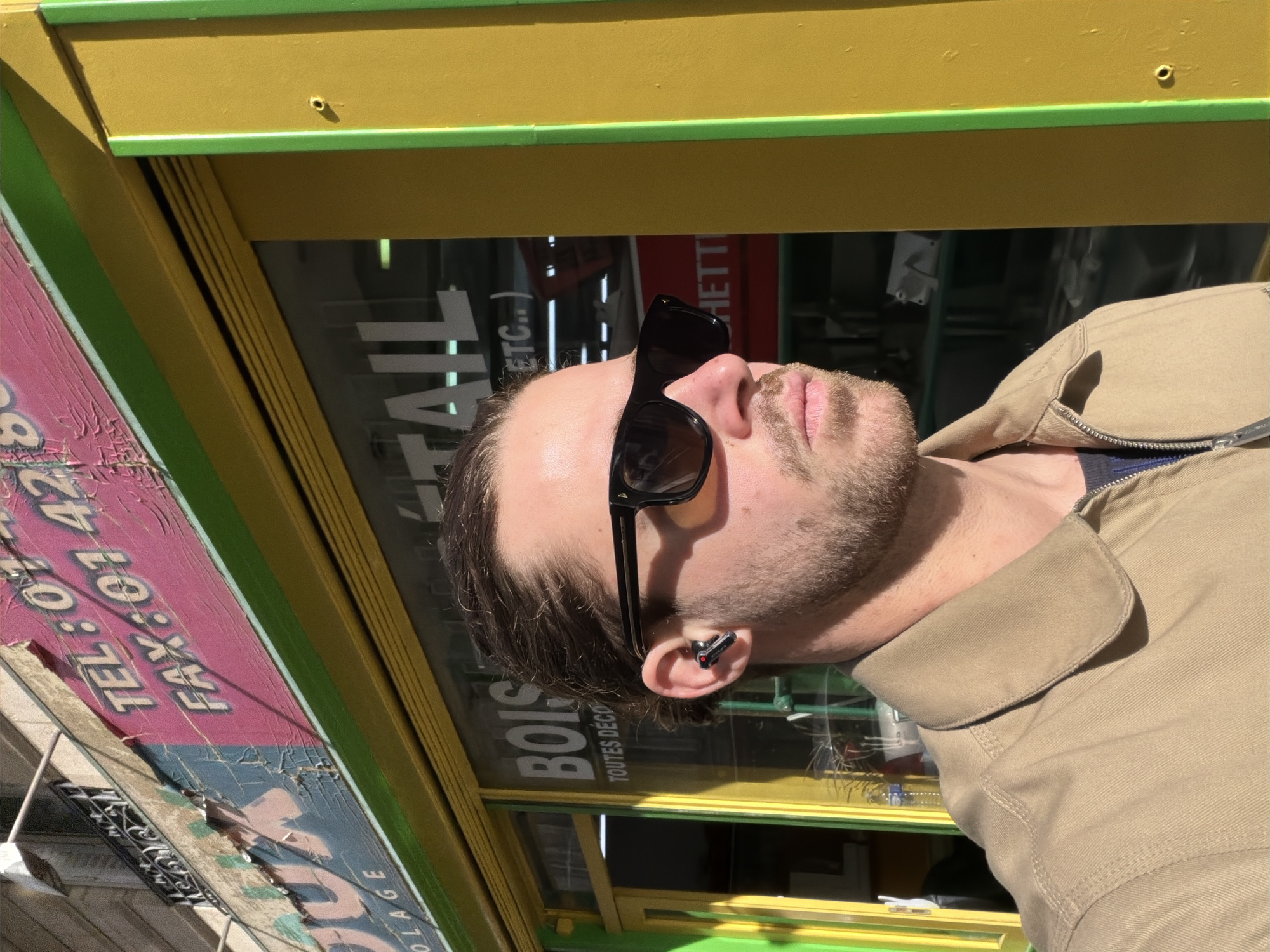 A man wearing sunglasses and an earpiece stands in front of a shop in Paris, France. The shop has a yellow and green exterior with pink signage on the left. Text visible on the signage includes telephone and fax numbers. The shop window contains the words "BOIS" and "TAIL". The man is wearing a tan jacket.