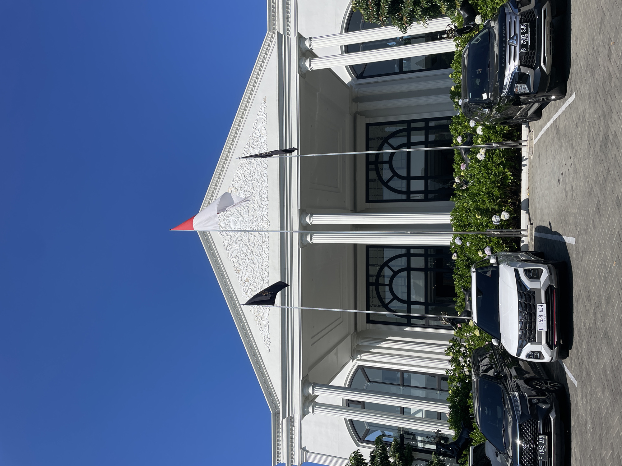 The image displays the exterior of a large, white building with neoclassical architectural features, located in Kayuambon, Indonesia. The building features a prominent triangular pediment adorned with intricate white bas-relief ornamentation, supported by several fluted white columns. Large arched windows or doors with black frames are visible on the facade.
Three flags are displayed on flagpoles in front of the building. The central flag is a red and white horizontal bicolour, positioned slightly higher than the two flanking flags. To its left and right are solid black flags.
In the foreground, a paved parking lot with interlocking brick-like pavers accommodates three parked cars. From left to right:
1. A black Toyota Veloz, with license plate F 1042 AAO, is parked facing the building.
2. A white Hyundai Creta, with license plate D 1598 AJH, is parked facing the building.
3. A black Mitsubishi Pajero Sport, with license plate B 2502 SJG, is parked facing the building.
All cars are positioned within marked parking spaces.
A line of dense green bushes interspersed with white and light purple flowers runs along the base of the building, separating the parking area from the structure. Partially obscured within these bushes are dark-colored decorative statues resembling deer or similar animals. The sky is clear and deep blue, indicating bright, sunny weather.