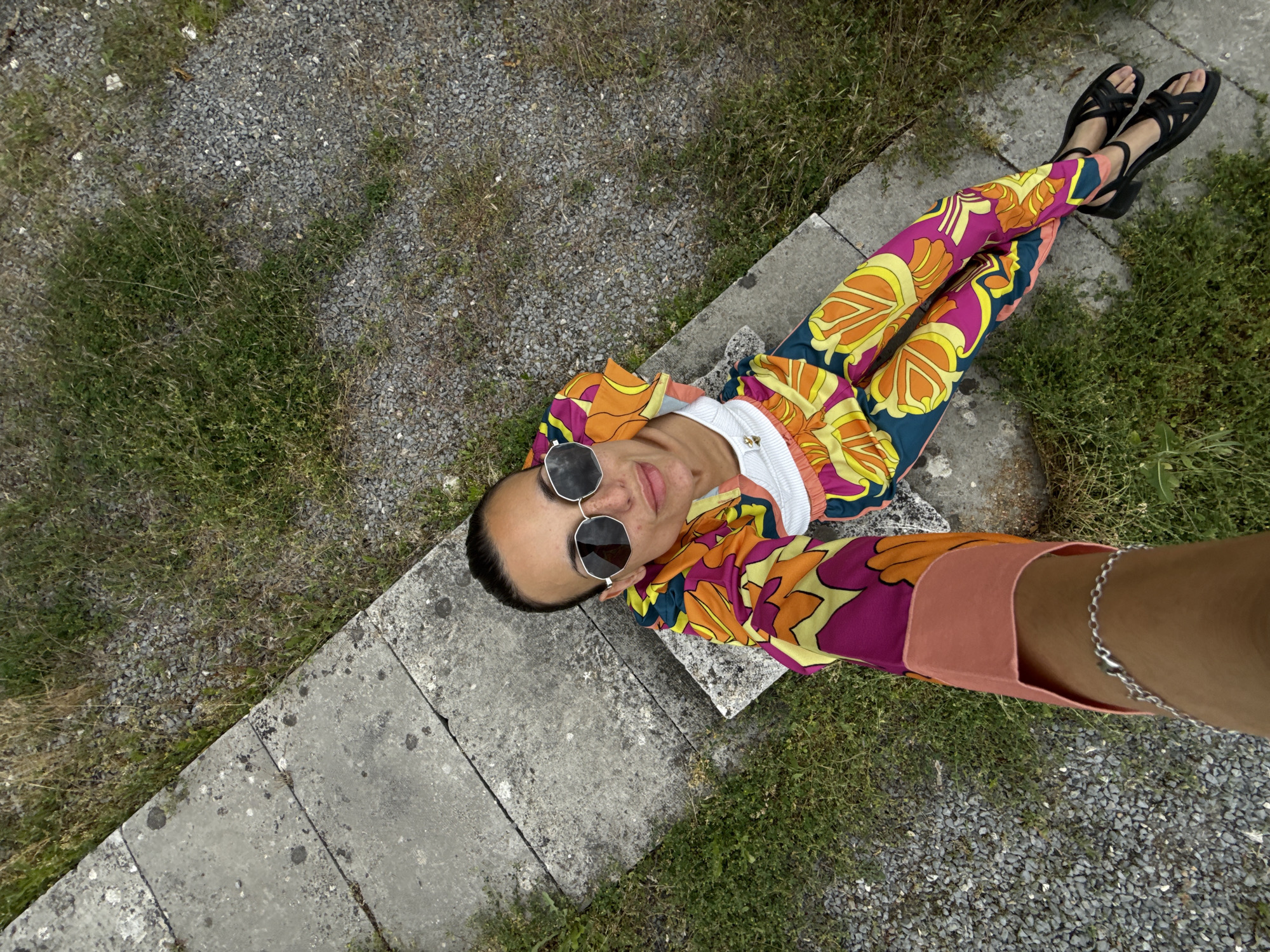 A high-angle, medium-shot selfie shows a woman sitting on a concrete structure outdoors. She is wearing sunglasses, a white top, and colorful patterned pants and a matching jacket. Her left arm is extended as if taking the photo. The background consists of gravel and grass. This image was taken at the Pantheon of Revivalists in Pleven, Bulgaria.