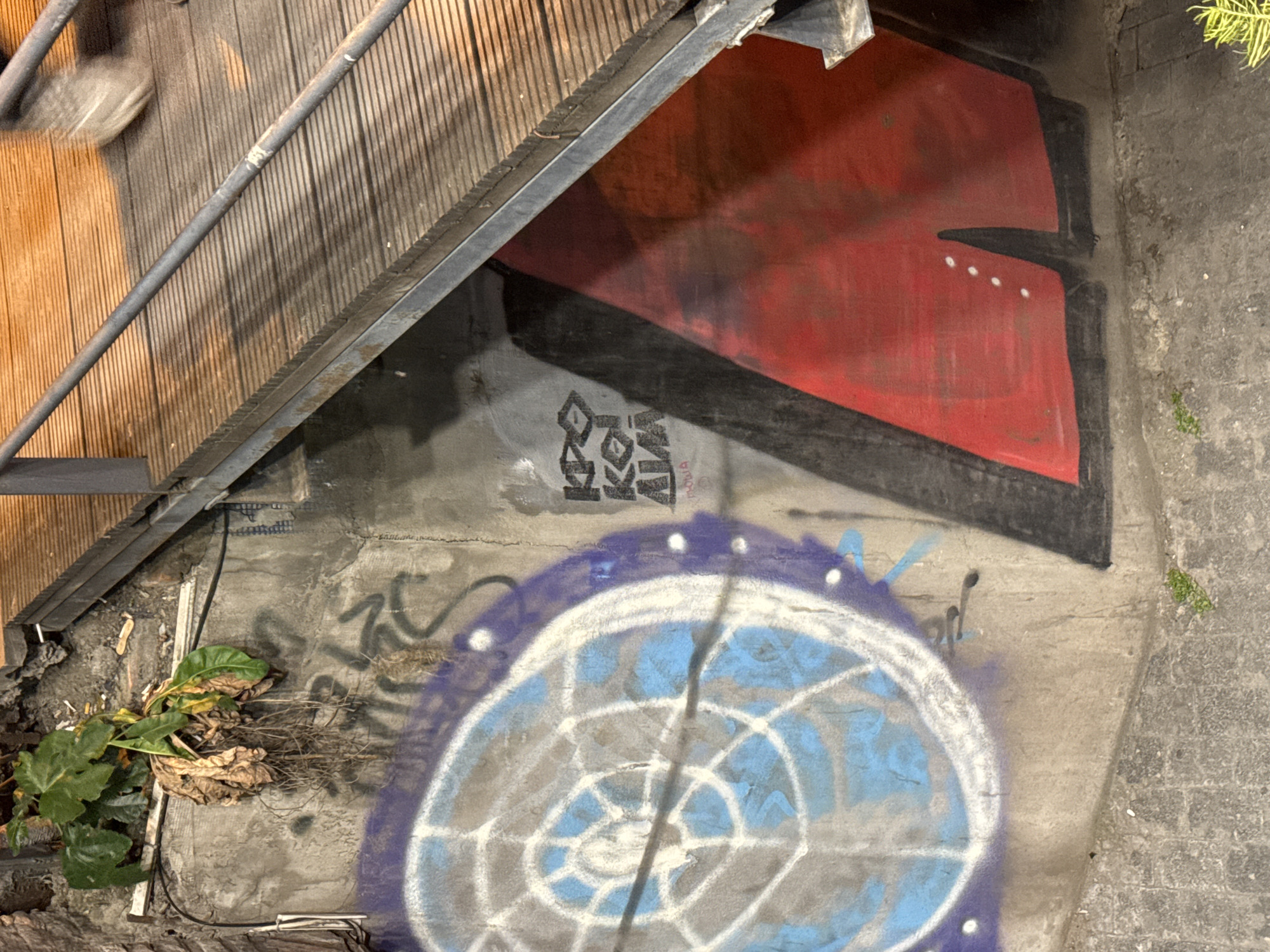 Outdoor urban scene in Tbilisi, Georgia. A concrete wall with graffiti, including a large red and black shape and a blue and white concentric circle design, sits below a metal staircase. Some plants and debris are visible at the wall's base. The ground next to the wall appears to be made of grey stone.