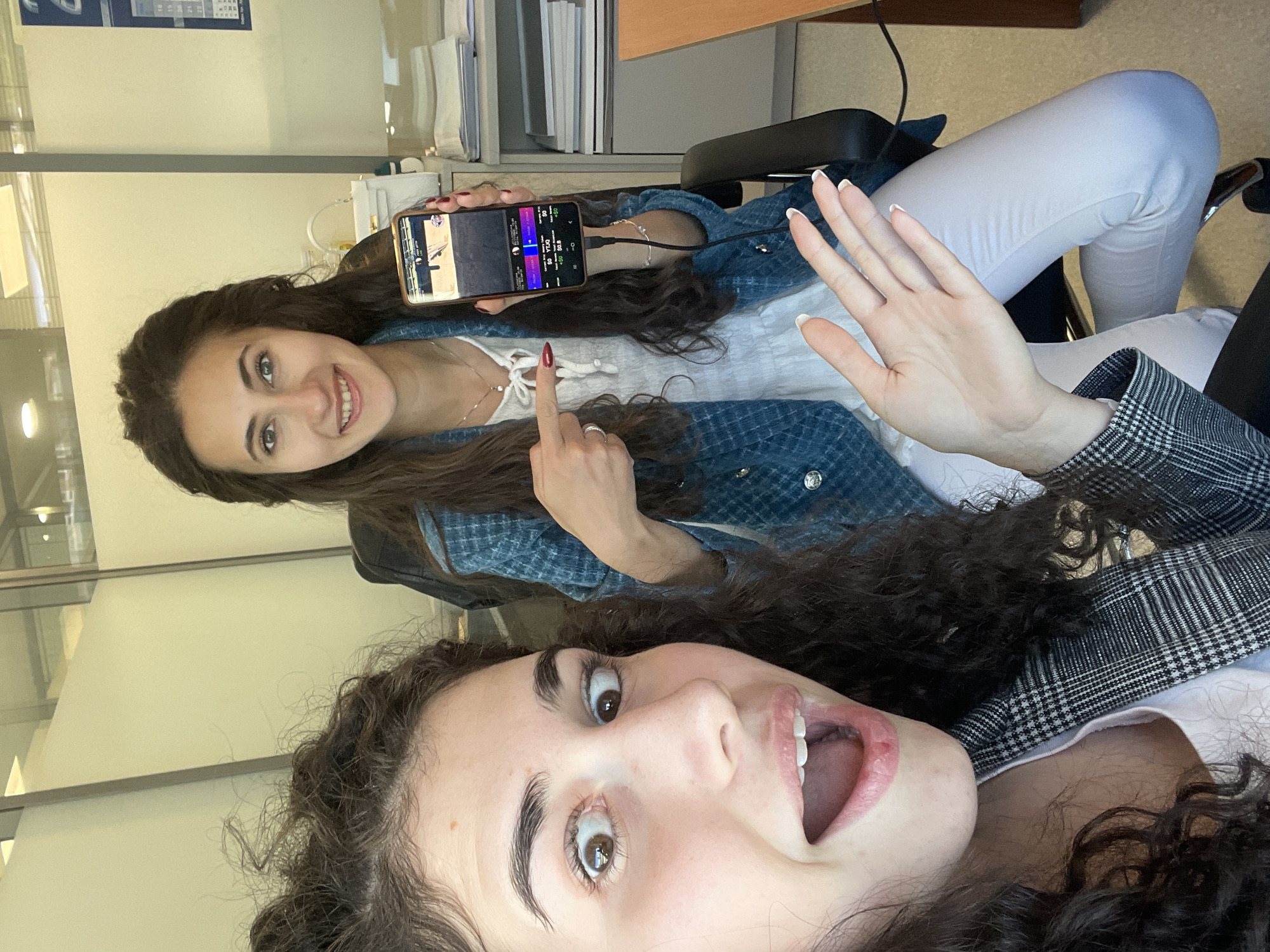 Two young women are posing for a selfie indoors. The woman in the foreground has curly dark hair and is smiling with her mouth open. She is wearing a white shirt and a black and white checkered jacket. The woman in the background has long dark hair and is smiling while holding a smartphone displaying what appears to be a sports betting interface. She is wearing a white top underneath a denim jacket. She is pointing with one finger towards the phone screen while her other hand is raised. They are both sitting in office chairs with white pants. Behind them is an office environment with desks, files, and glass partitions. The precise location is unknown and there's no information indicating the city of Sofia, Bulgaria.
