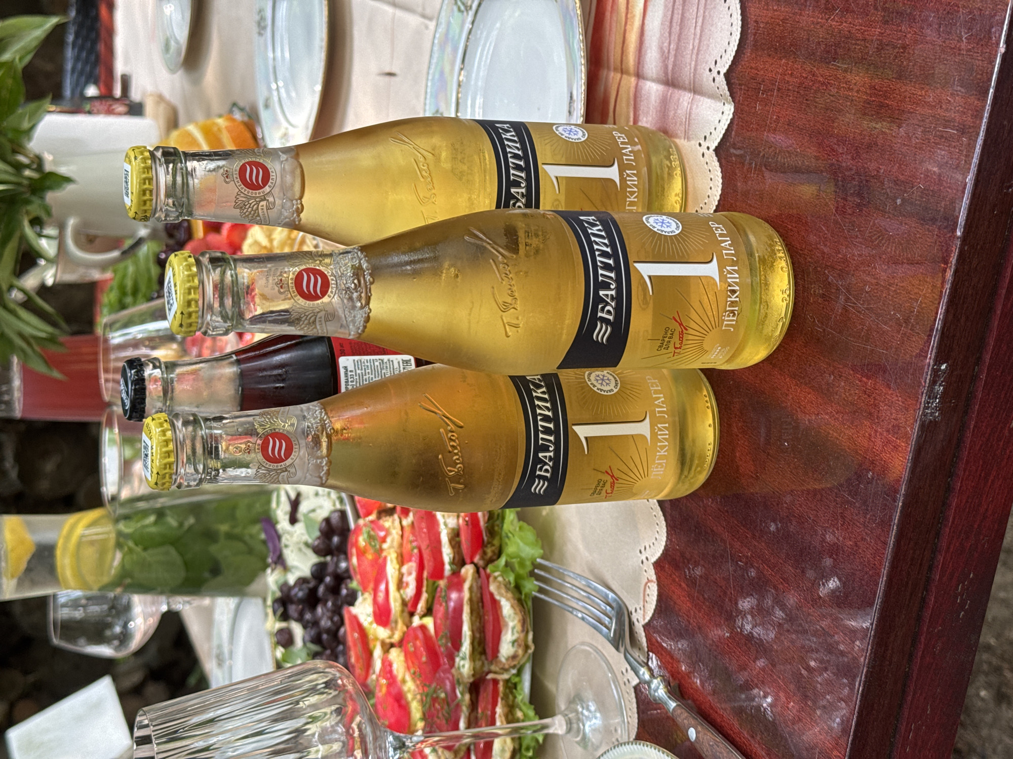 The image depicts an outdoor gathering on a wooden table, likely in Буран Снт, Russia, as indicated by the context. Three bottles of "Baltika 1" light lager are prominently displayed in the foreground. The bottles are golden yellow, filled with a pale yellow liquid, and have yellow caps with red and white labels. The labels feature the brand name "Балтика," the number "1," and text that reads "лёгкий лагер" (light lager).
In the background, a platter of food is visible, featuring circular slices of food topped with red tomato slices and green herbs, possibly appetizers or small dishes. There are also glasses on the table, including a wine glass on the left and a tall glass with what appears to be water and mint. Other elements in the background include a white pitcher or jug, some greenery from a plant, and parts of plates. The overall setting suggests a relaxed outdoor meal or social event. The lighting indicates it is daytime, with natural light illuminating the scene.