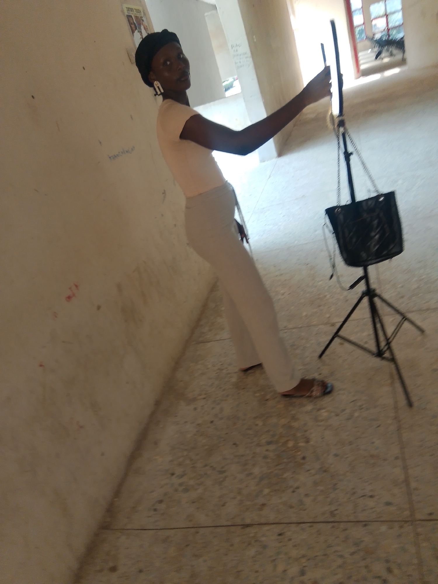 In the city of Jalingo, Nigeria, a young Black woman is captured in a hallway. She is wearing a black headscarf, a light peach t-shirt, and light-colored trousers. Large, white, geometric earrings adorn her ears. She is holding a thin, black vertical object, possibly a light ring or a camera accessory, in her right hand. Her gaze is directed towards the camera.
In the background, a black handbag hangs from a tripod. The hallway walls are beige and bear some faint markings and scribbles. The floor is a speckled gray concrete. Sunlight streams in from an opening at the end of the hallway, illuminating the scene. The overall impression is one of an indoor setting, perhaps a school or a public building, with the woman actively engaged in what appears to be content creation or a similar activity.