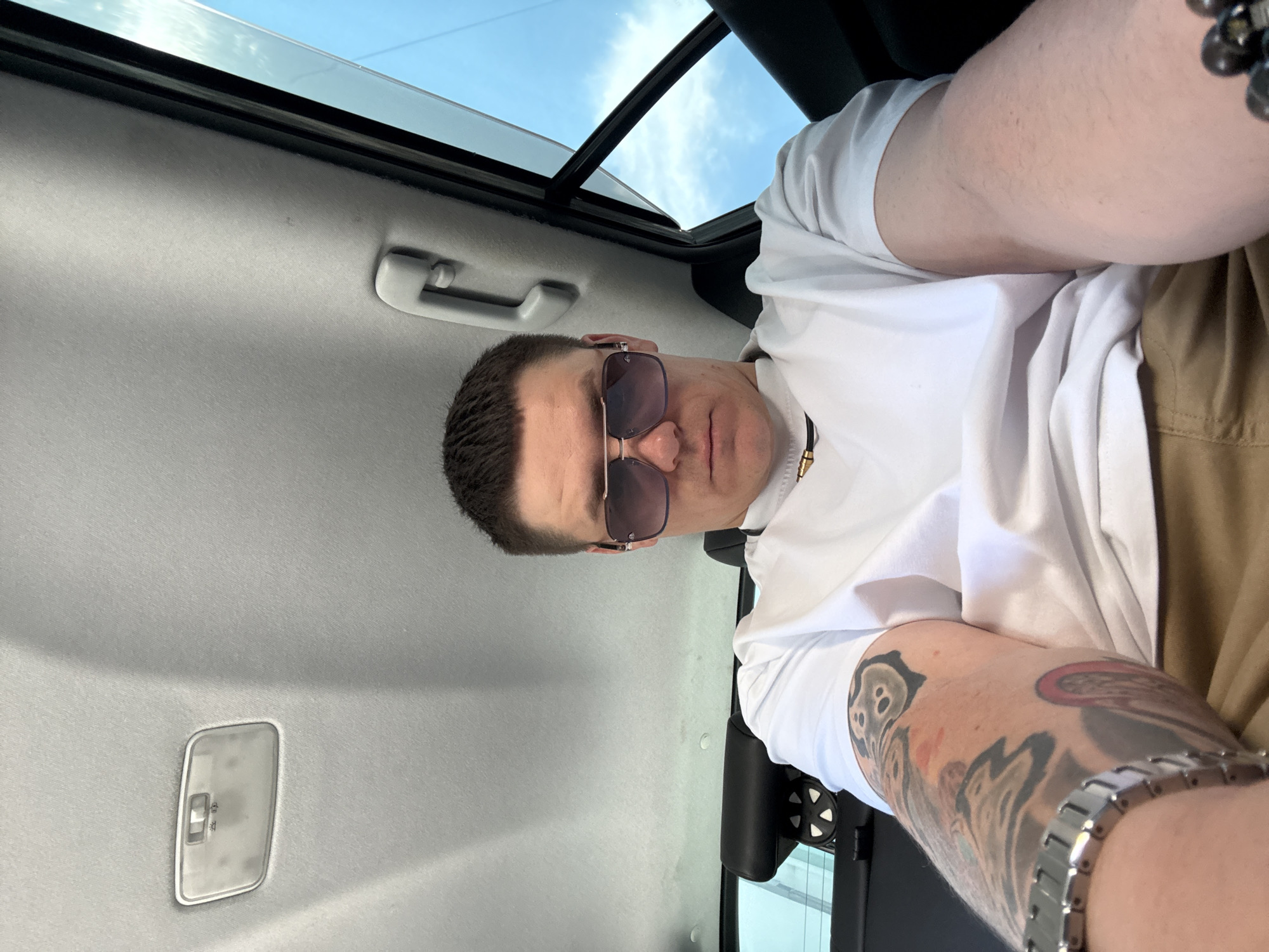 A man with short dark hair and sunglasses is sitting in the back seat of a car. He is wearing a white t-shirt and khaki pants. He has tattoos on his left arm and is wearing a silver watch. The car's interior is visible, including the ceiling light and a grab handle. Outside the car window, the sky is visible. The image was taken in Vladivostok, Russia.
