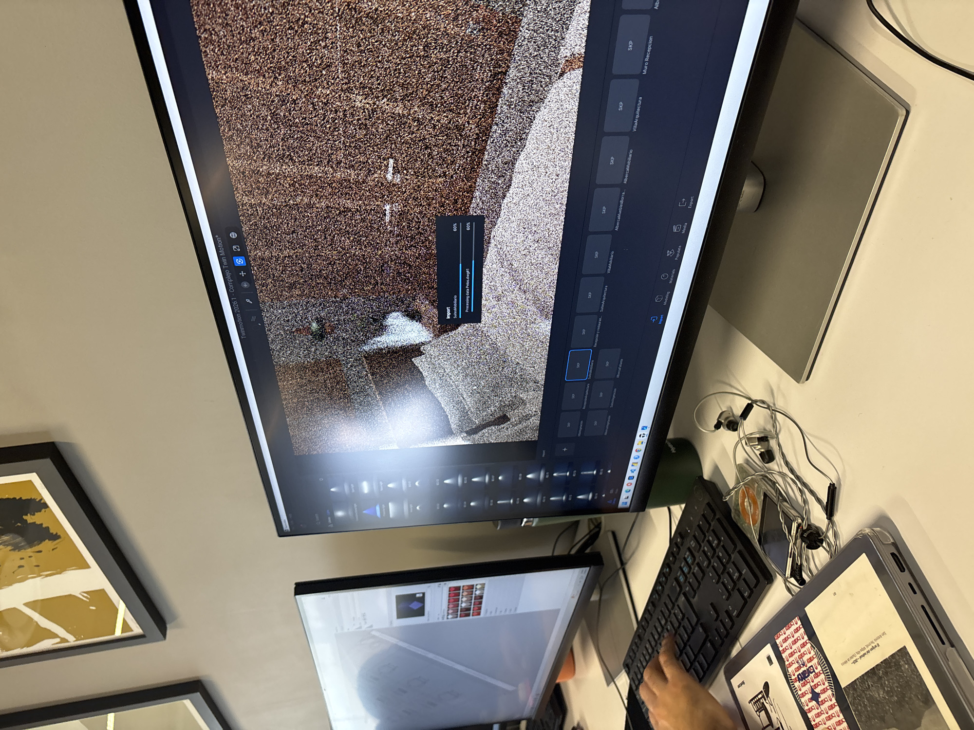 Here is a description of the image. The scene shows a desk with two computer monitors, a keyboard, a tablet in a gray case, and earphones with a phone underneath. The top monitor displays architectural software, possibly Twinmotion, showing a 3D model being imported, with the import progress at 60%. The secondary monitor below shows another software with a photo editing-like interface. On the tablet, there is an open book, featuring text in Spanish. Also present are framed pictures on the wall, suggesting an office or home office setting. Based on the OCR text on the screen "San Antonio Tin Mpa Atau," it indicates a possible location of this Twinmotion project in Mexico City, Mexico.
