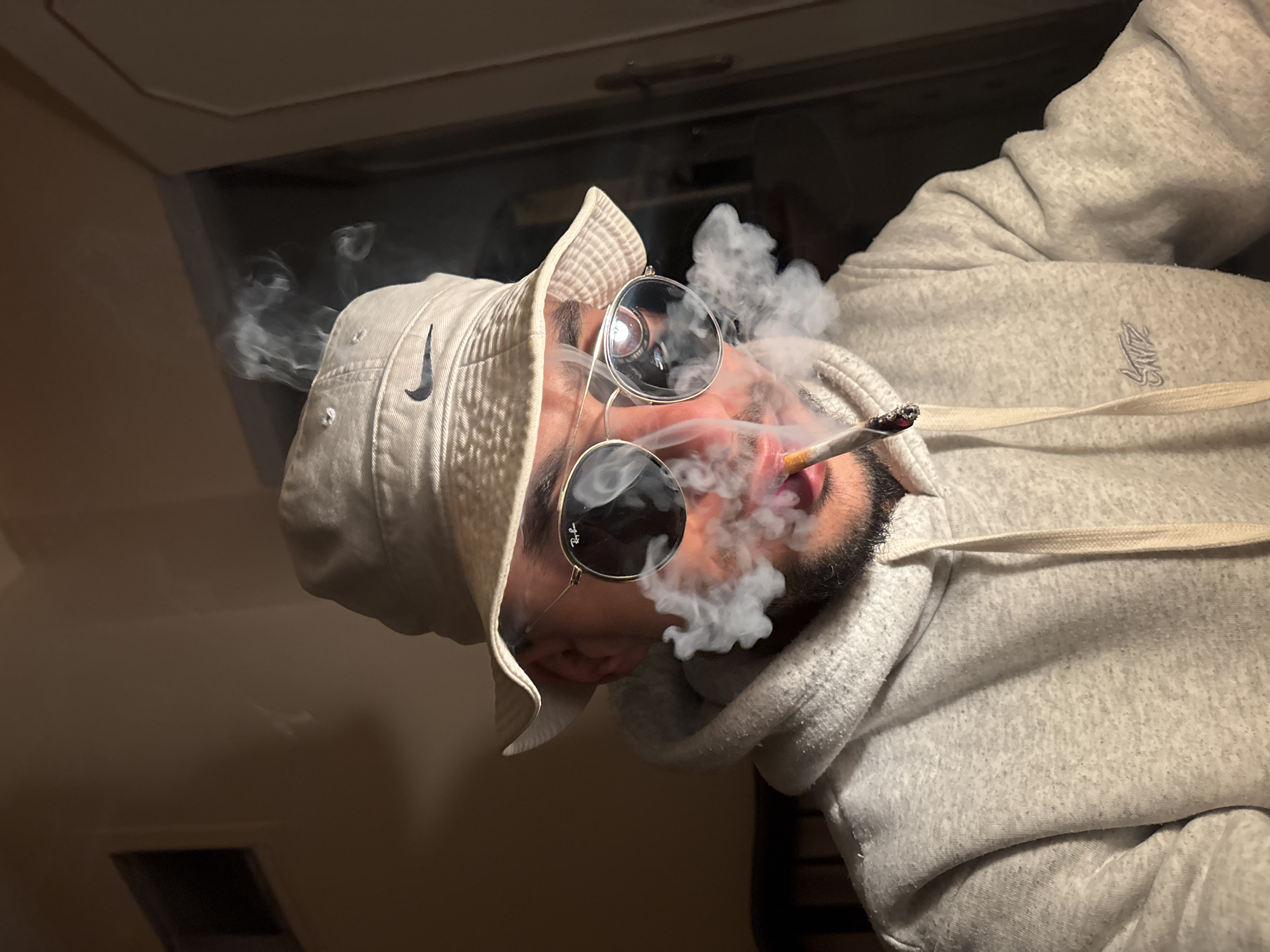An individual is depicted indoors in Tanger, Morocco, in a low-light environment. The subject, with visible beard stubble, wears a beige Nike-branded bucket hat, round gold-rimmed Ray-Ban sunglasses, and a light gray hooded sweatshirt bearing a 'SNTZ' emblem on the left chest. A lit, hand-rolled cigarette is positioned between the subject's lips, from which dense white smoke is actively expelled, partially obscuring the lower face. Reflections of an apparent light source are visible in the sunglasses. The indoor background consists of a light brown wall featuring a dark rectangular ventilation opening on the upper left, and a partial white door frame with a visible handle on the right. Dark, slatted furniture is visible in the lower left portion of the image.