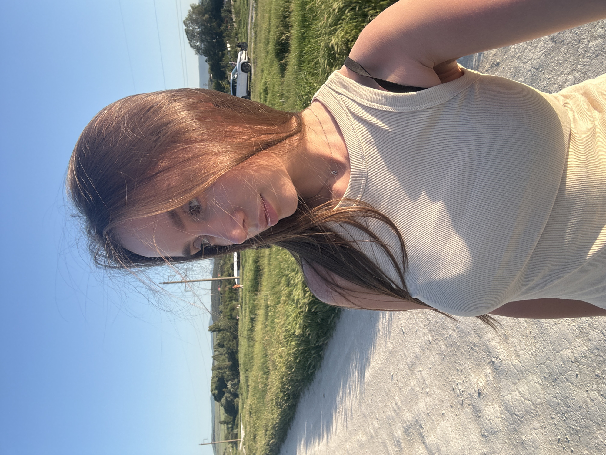 A young woman stands on a light-colored dirt path in Вишенское сельское поселение, Ukraine. She wears a light-colored ribbed tank top and a delicate necklace. Her hair is long and brown. A grassy field stretches out beside the path, and in the distance, there is a parked white vehicle. Overhead utility lines stretch across the clear blue sky.