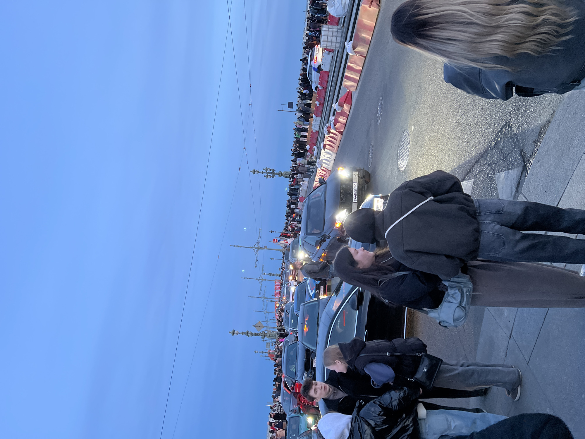 The image shows a crowd of people on a bridge in Saint Petersburg, Russia, likely during a public event. Cars are stationary on the bridge. Several people are standing on the pedestrian walkway beside the road. The blue sky suggests early evening light. A long line of vehicles is stopped on the bridge, some with headlights on. Barriers delineate the traffic lanes and pedestrian areas. The license plate E440OE 155 is visible on a car.
