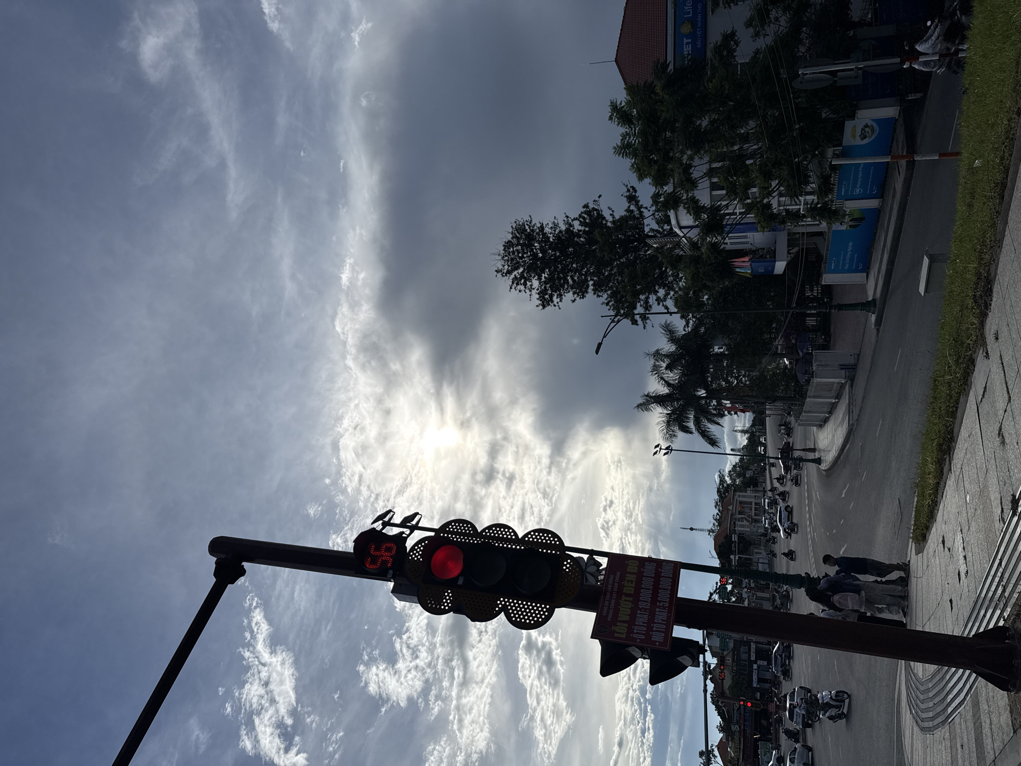 The image depicts a street scene in Da Lat, Vietnam, featuring a traffic light with a red signal and a countdown timer displaying "56". The traffic light is positioned on a pole alongside a sidewalk with textured paving. Below the traffic light, a red and white sign with Vietnamese text is visible, seemingly indicating traffic violations and associated fines.
In the background, a street intersection is busy with traffic, including numerous motorbikes and a few cars. Several buildings line the street, some with awnings and signs, one of which appears to be a commercial establishment with a blue logo and English text "NET Life". Palm trees and other greenery are interspersed throughout the scene, contributing to the tropical urban environment. The sky above is partly cloudy with bright sun peeking through, casting dramatic shadows and light patterns on the scene. A group of four individuals, two adults and two children, are standing on the sidewalk near the traffic light, observing the traffic or waiting.