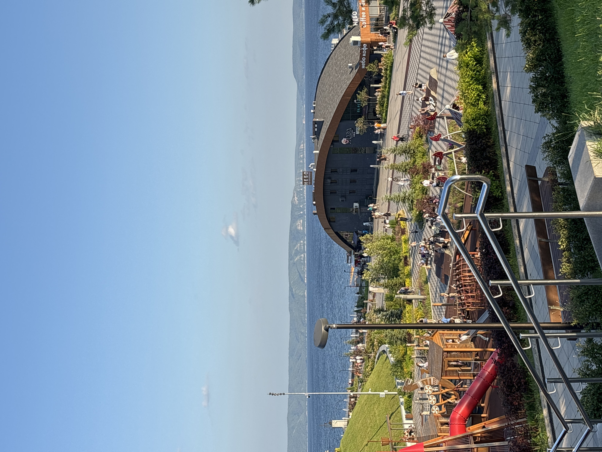 The image depicts a sunny day at a waterfront park in Tolyatti, Russia, likely on the banks of the Volga River. In the foreground, a metal railing guides a downward sloping path. To the left, a wooden playground structure with a red slide is visible, accompanied by various children's play equipment. Green grass and bushes line the pathways.
In the middle ground, a large, modern building with a curved, dark-tiled roof dominates the scene. Signs on the building indicate it houses establishments like "НЕБО ГАСТРОЦЕНТР" (Nebo Gastropoint) and "НЕБО coffee и...". People are seen walking and congregating in front of and around this building, suggesting it is a public space with amenities. A paved plaza with a patterned design is in front of the building.
Beyond the building, a wide, calm body of water, likely the Volga River, stretches to the horizon. Distant, rolling green hills form the opposite shoreline. The sky is a clear, bright blue with a few wispy clouds. The overall atmosphere is one of a recreational area with families and individuals enjoying outdoor activities.