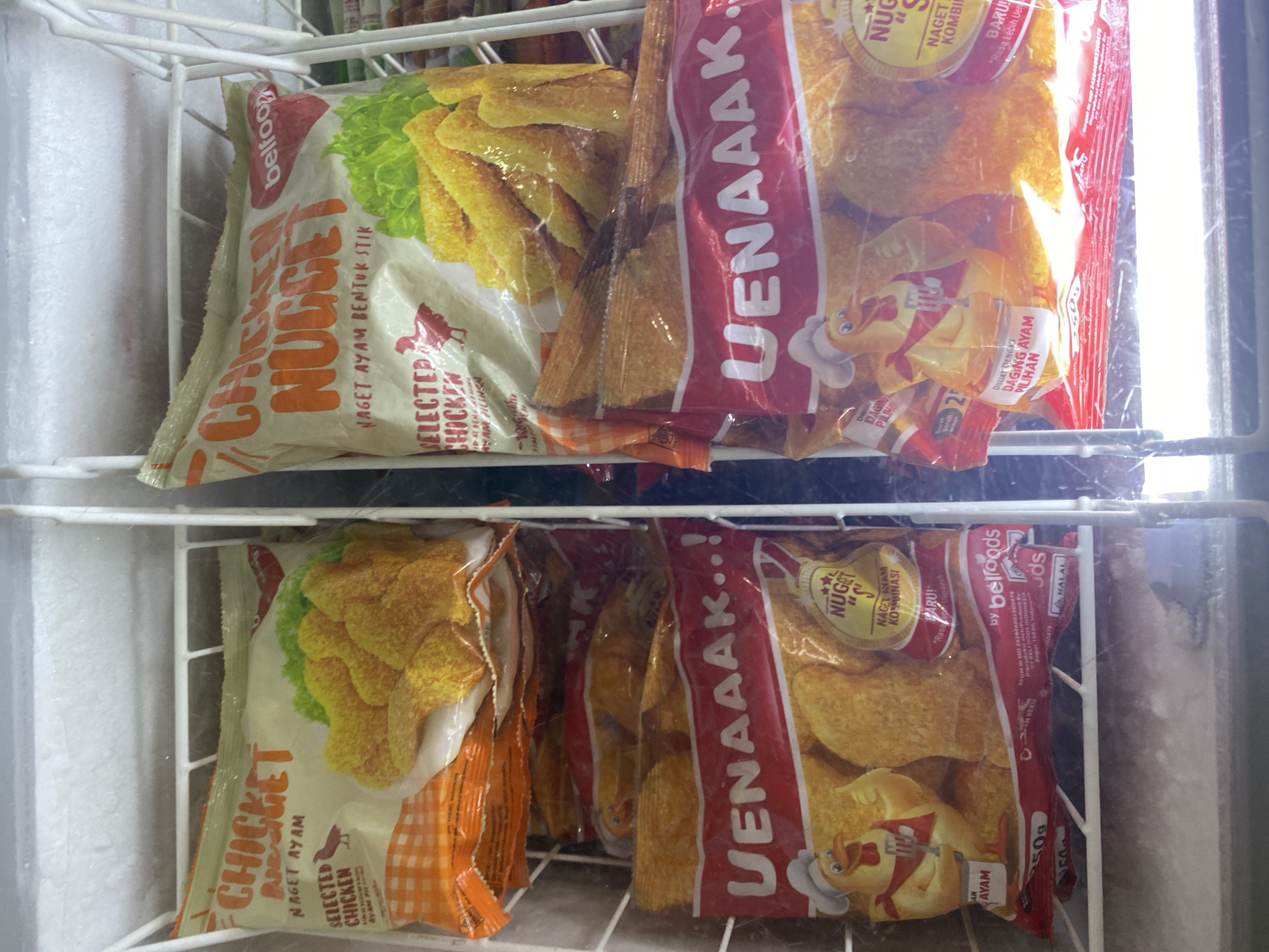 The image shows a freezer filled with frozen chicken nugget products. Two brands are visible: "CHICKET NUGGET" and "CHICKEN NUGGET". Both appear to be packaged in colorful plastic bags with images of chicken nuggets and other branding elements. The products are arranged in compartments within the freezer. The lighting is cool and artificial, consistent with a freezer environment. There are no people or animals present in the image, and no visible interaction is occurring. The location of Payatieng, Indonesia is not evident in the image itself, as there are no identifying landmarks or signs related to that specific city. The image is a close-up view of the frozen food items within the freezer.