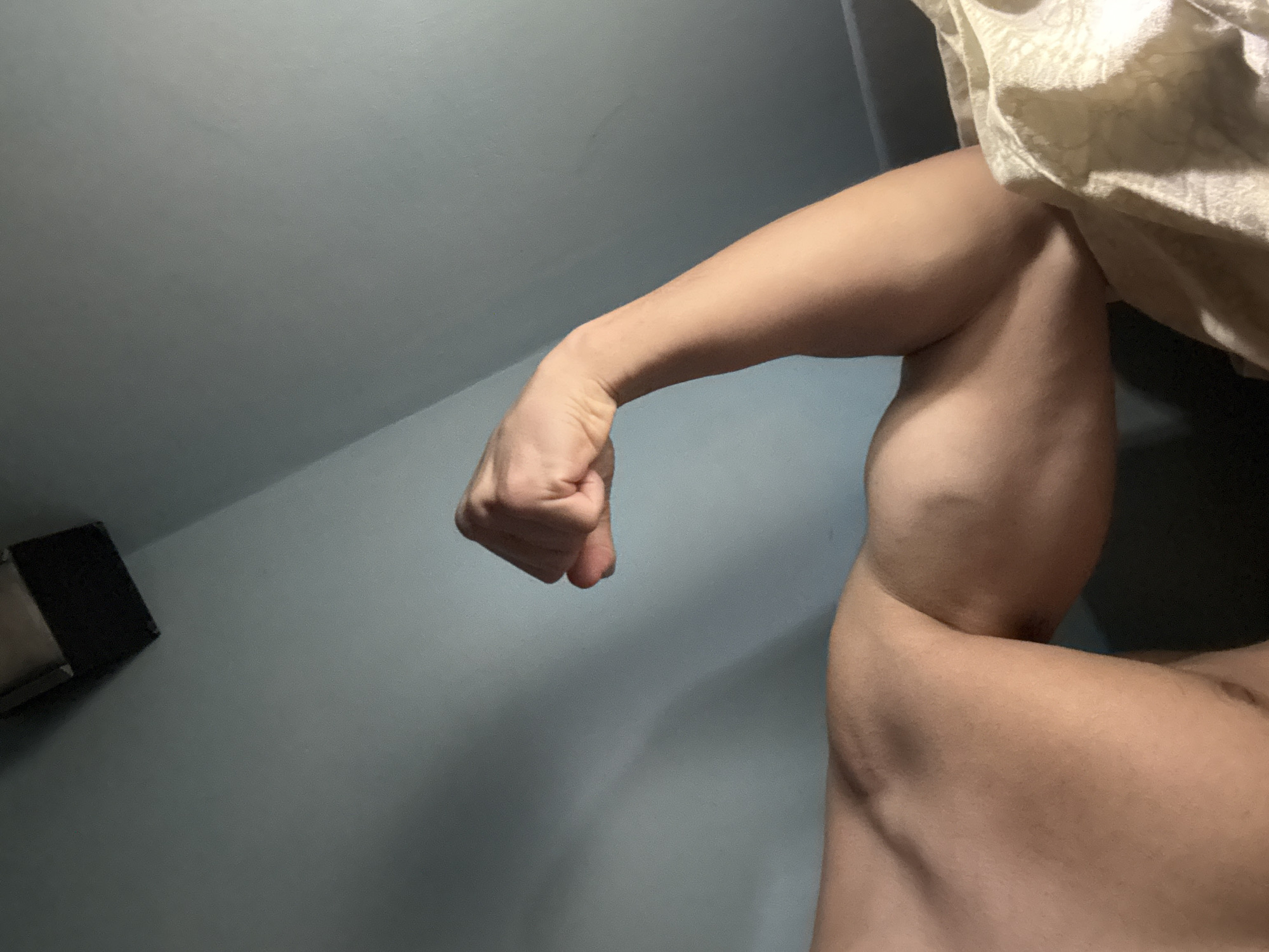 The image shows a muscular arm flexing its biceps against a blue-gray wall. A partially obscured off-white fabric is present in the upper right corner. There is a black object in the lower left corner of the image. There is no information to determine the location is in Buhovo, Bulgaria.