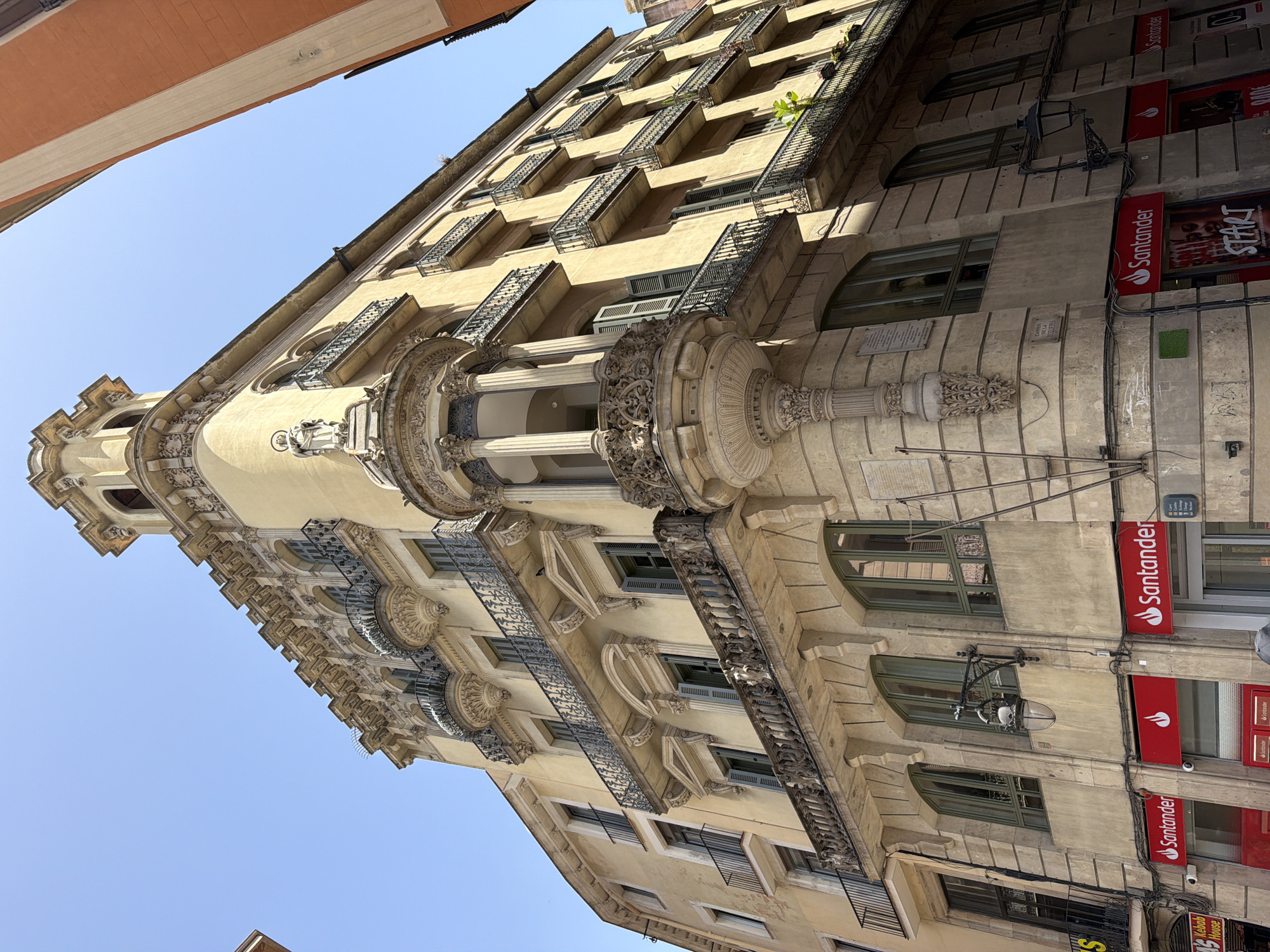 This is a low-angle, outdoor, eye-level shot of a multi-story building, likely in Barcelona, Spain, as indicated by a street sign that reads "CARRER DE LA BOQUERIA". The building is ornate, with a yellow facade, decorative balconies, columns, and intricate stonework. The sun is shining brightly, casting shadows and illuminating the architecture.
The building appears to house several businesses on its ground floor, with prominent red signs for "Santander" visible. Another sign displays the word "START", and a smaller sign indicates "Kebab House". The upper floors feature numerous windows and balconies with dark metal railings, some adorned with small plants. One balcony on a lower, more prominent section of the building is particularly elaborate, featuring detailed sculptures and columns.
There are no people clearly visible in the image, suggesting the photo was taken during a time when the street was not heavily populated or the focus is solely on the architecture. The sky is a clear, bright blue, indicating fair weather. The overall impression is a blend of historical architecture and modern commercial activity.