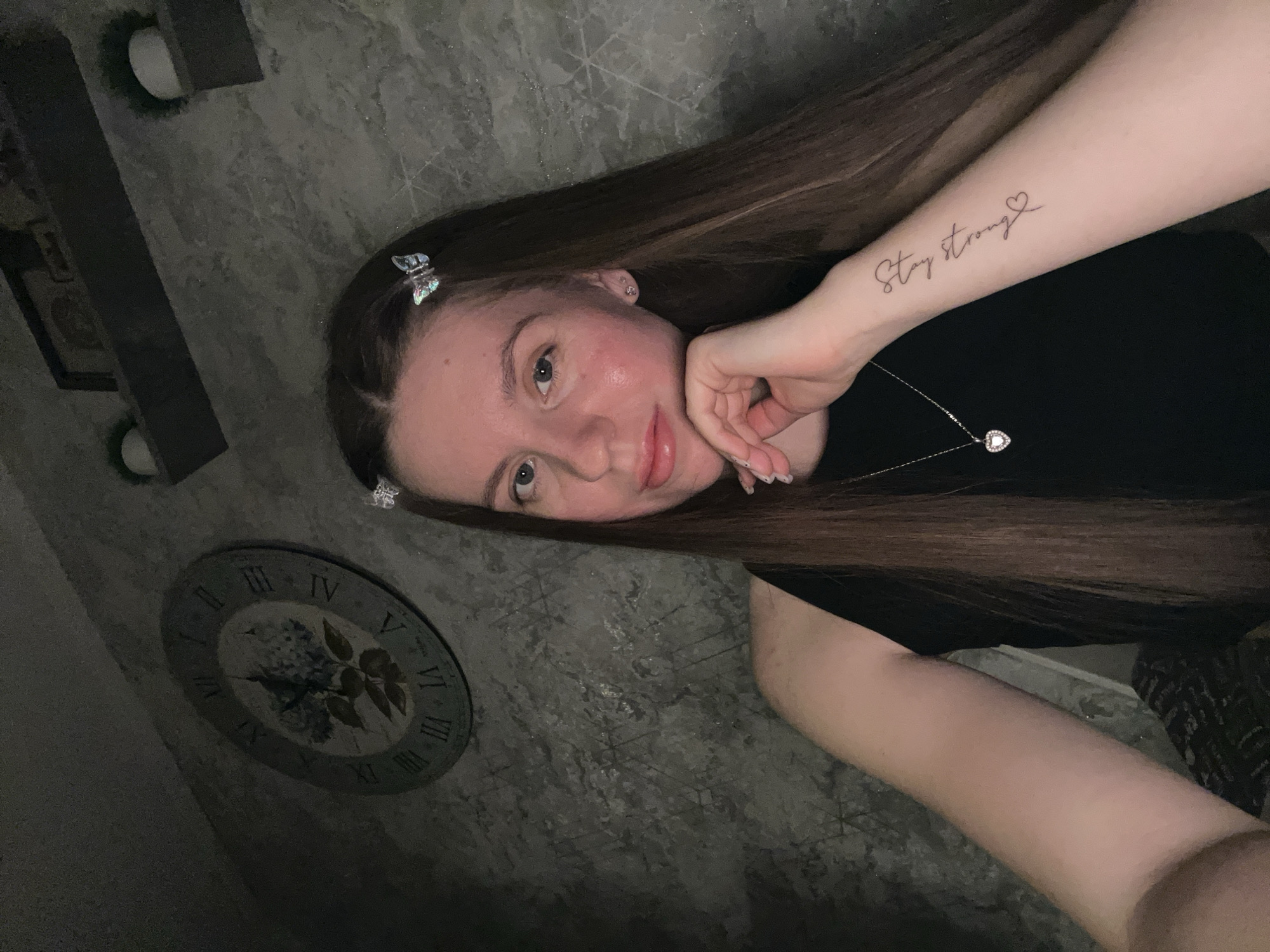 A young woman with long brown hair is reclining in what appears to be a dimly lit indoor setting. She is wearing a black top and a silver necklace with a heart pendant. Her left forearm is visible, bearing a tattoo that reads "Stay Strong" with a small heart design. Her hair is adorned with butterfly clips. A clock is visible on the wall in the background, and a patterned wall texture is present. The location in the city of Klin, Russia is not verifiable from the image.