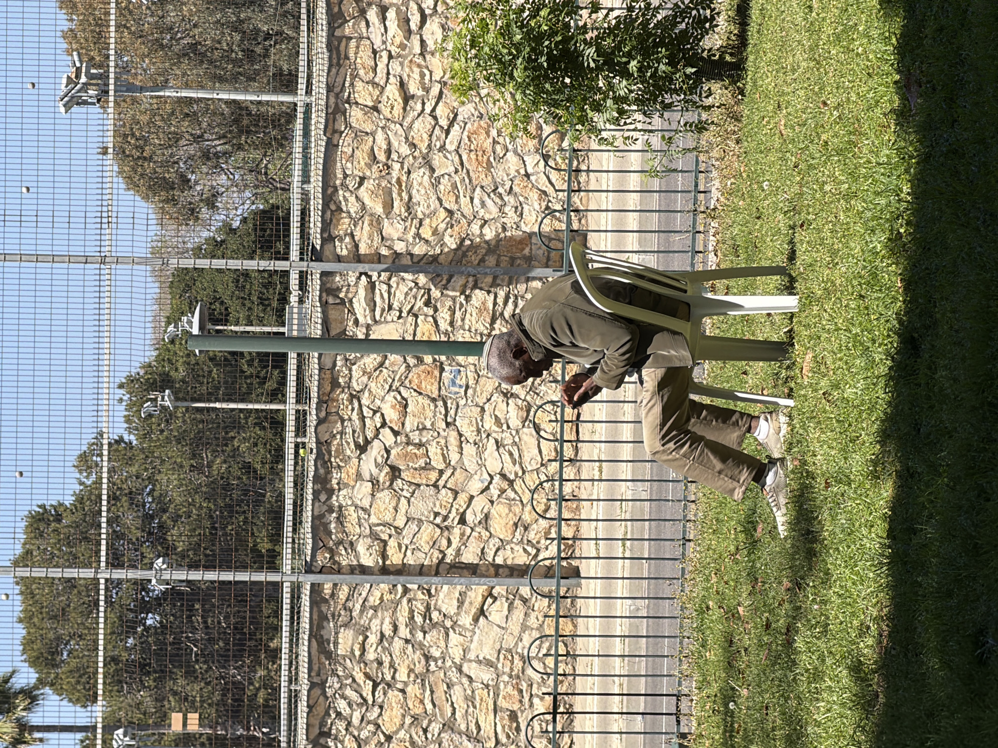 An outdoor scene captured during bright daylight shows an older man seated on a light-colored plastic chair, looking downwards, possibly at an object in his hands. He wears a dark olive green long-sleeved top, brown or olive green pants, and light-colored sneakers. The man is situated on a lush green lawn, with a paved walkway visible to his left.
Behind the man, a dark metal railing borders a tall metal mesh fence, which encloses what appears to be a sports court, possibly for tennis or padel. Within the court area, several large trees with dense green foliage are visible under a clear blue sky. Two light-colored balls, consistent with tennis or padel balls, are suspended in the air near the fence, suggesting an ongoing or recently completed game. Metal poles fitted with lighting fixtures or cameras are also present along the fence line.
Beyond the sports court fence is a substantial wall constructed from irregularly shaped, light-colored stones. A small, leafy green bush grows above this stone wall. Bright sunlight illuminates the scene, casting distinct shadows on the grass and the stone wall, indicating a sunny day in Tel-Aviv, Israel. The man appears to be resting or observing the surroundings in this recreational setting. No visible text is present.