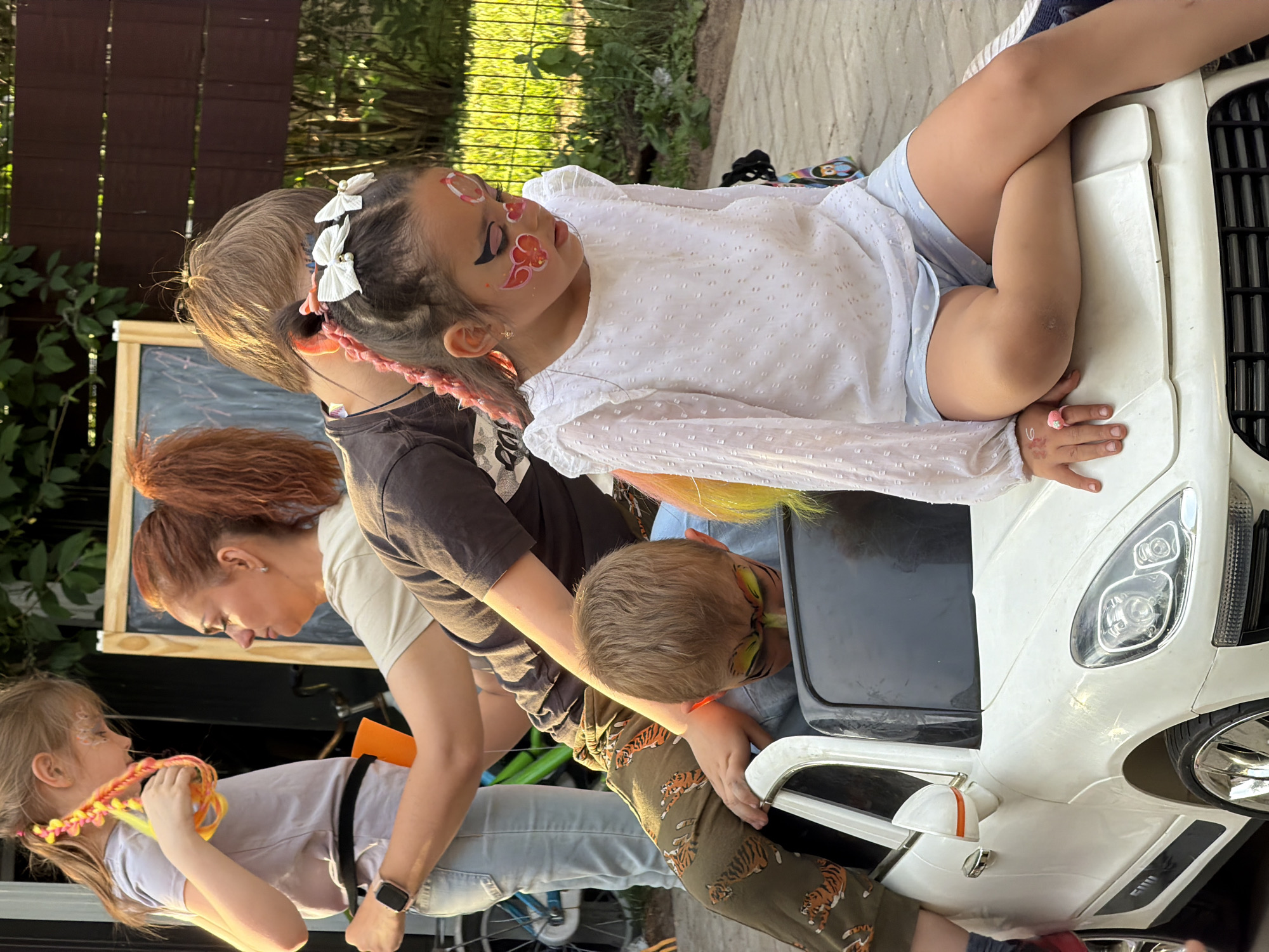 The image depicts an outdoor children's gathering, likely a party or playdate, bathed in the warm, late afternoon or early evening sunlight, suggesting a clear and sunny day in Riga, Latvia.
In the foreground, a young girl is prominently featured, leaning on a white ride-on toy car. She has intricate face paint with red floral designs and black accents around her eyes, complemented by pink braided hair extensions and white bow hair clips. She wears a white long-sleeved top and light blue shorts.
Adjacent to her, a boy with short brown hair sports yellow, green, and black face paint, possibly a tiger design. He is dressed in olive green pants adorned with tiger patterns and appears to be interacting with the toy car. To the far left, another young girl with blonde hair, also with sparkly face paint on her cheek, holds colorful braided extensions. She wears a light purple t-shirt. An adult woman with auburn hair is leaning over, engaging with this blonde girl. A fourth child, wearing a dark t-shirt and with partially visible red/orange face paint, is also present behind the central girl.
The setting includes a wooden fence and lush green foliage in the background, indicative of a garden or backyard. A wooden easel or blackboard stand is visible to the left. A small backpack featuring a Mickey Mouse character can be seen near the toy car. The overall mood appears joyful and relaxed, with the children engaged in playful activities. No discernible text is visible in the image.