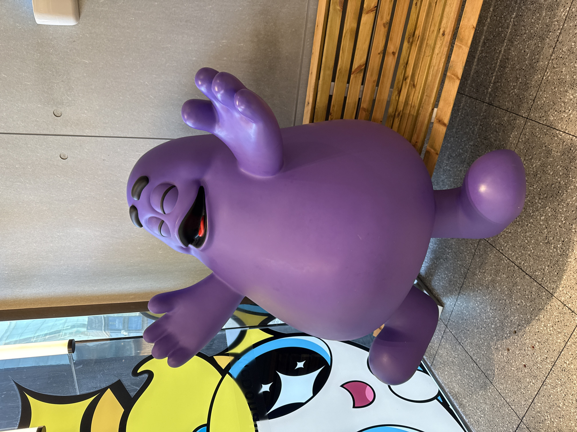 The image shows a purple, cartoonish figure with multiple eyes and an open mouth, posed with raised arms and a tail-like appendage. It appears to be a sculpture or statue and is standing on a surface decorated with cartoonish graphics. Behind the figure is a wall with wooden slats and tiled flooring. The image gives no indication of the location being in the city of Dongcheng District, China.