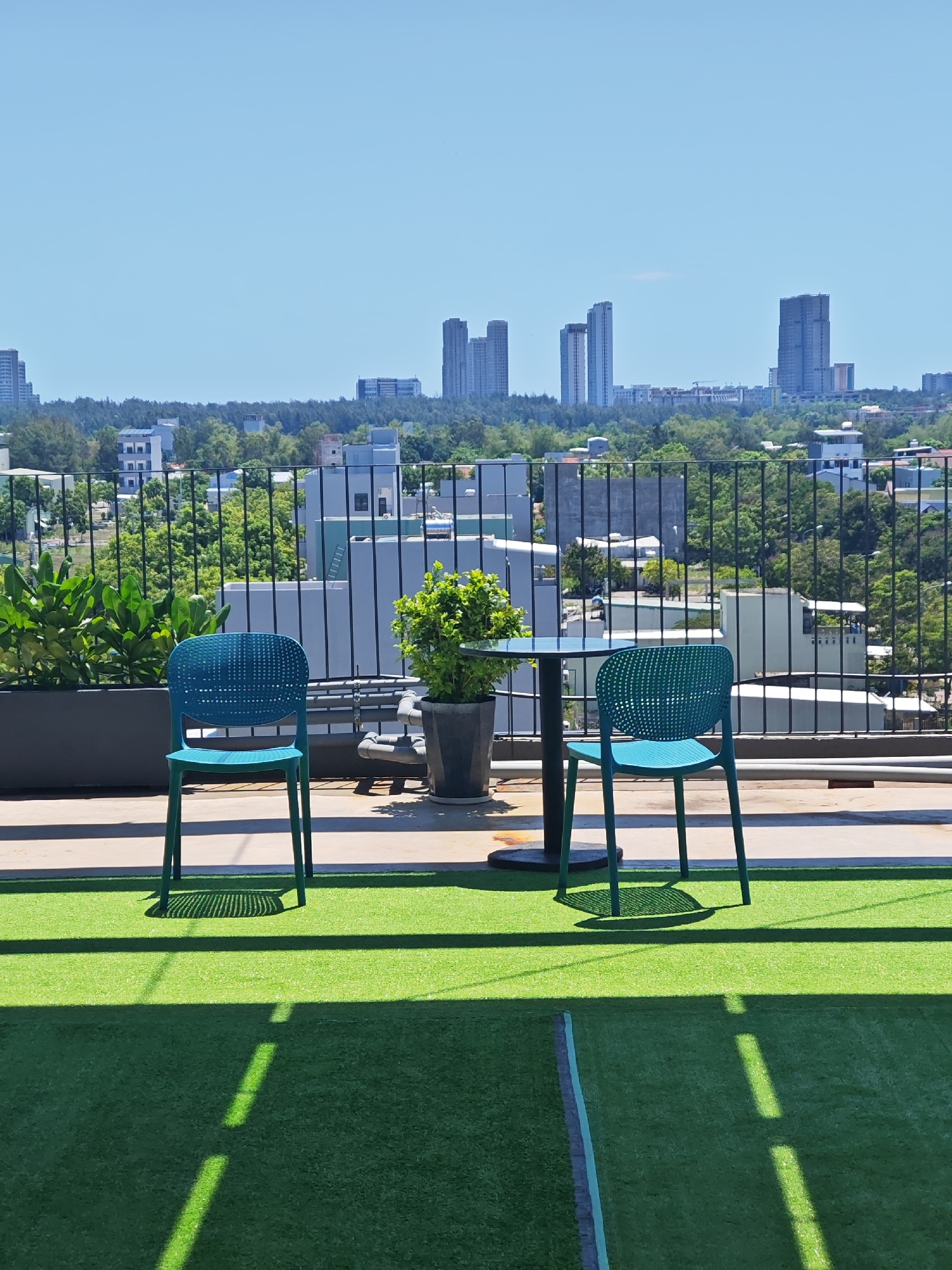This is an outdoor scene on a rooftop in Da Nang, Vietnam, with a clear blue sky above. In the foreground, an artificial green lawn covers the surface, with distinct shadows cast by an unseen structure. Two teal-colored chairs are positioned on the lawn, facing away from the viewer, with one chair slightly closer to a small, round table. The table is dark, and a potted plant with green foliage sits to its left.
Beyond the rooftop area, a black metal fence separates the immediate space from a wider view of the city. In the middle ground, a dense arrangement of green trees and various buildings, mostly light-colored, are visible. Some of these buildings are residential, with multiple windows, while others appear to be commercial or office spaces.
The background is dominated by a cityscape featuring several tall, modern skyscrapers. These buildings are gray and have a contemporary architectural style. The overall impression is a contemporary urban environment with recreational space on a rooftop overlooking the city.