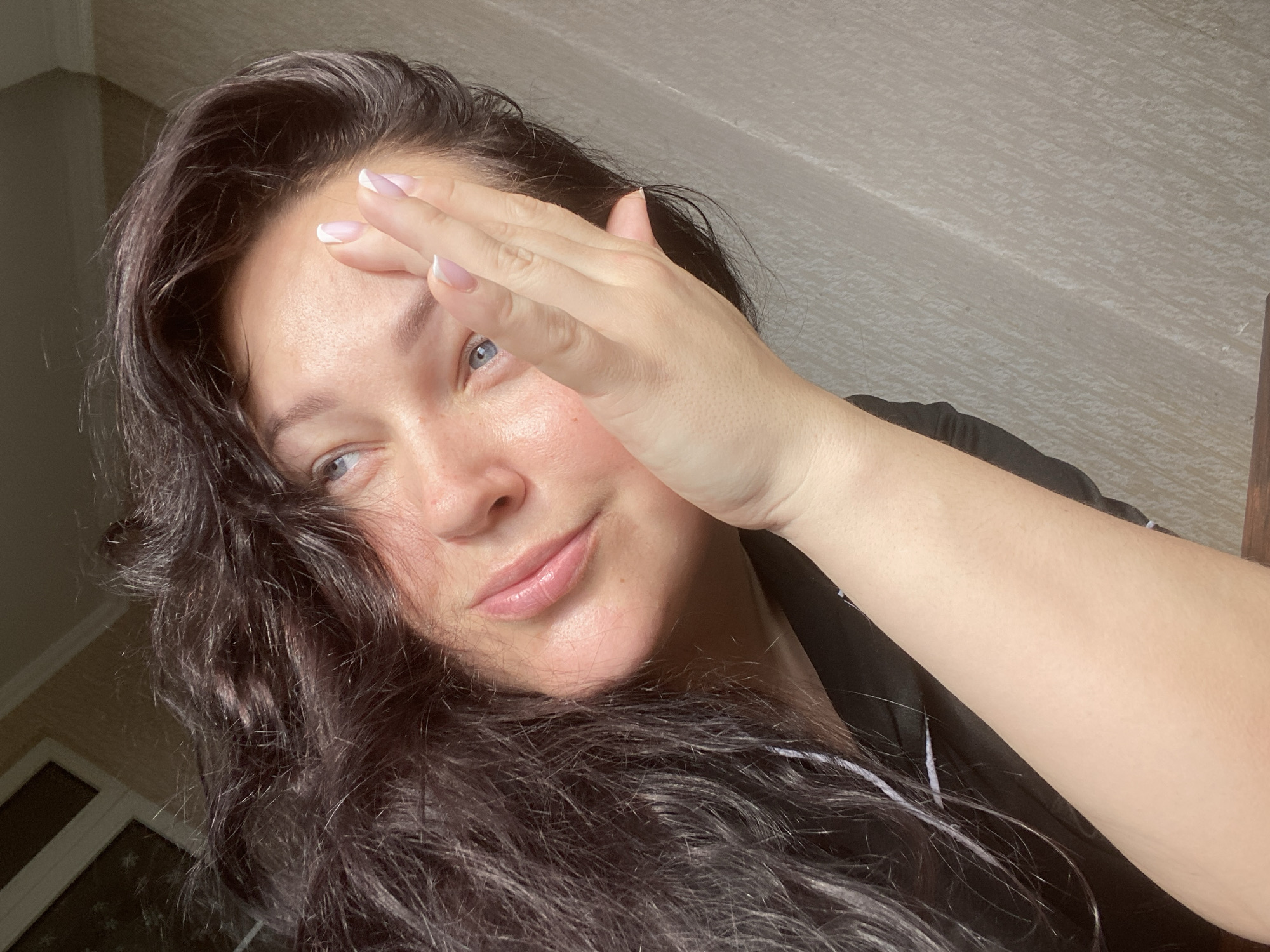 A woman with long, dark, wavy hair and light blue eyes is pictured in a selfie taken indoors. She is partially obscuring her forehead with her right hand, which features a French manicure. The background appears to be a textured wall. The location cannot be determined from this image, but given the instructions, it is supposedly in Sochi, Russia.