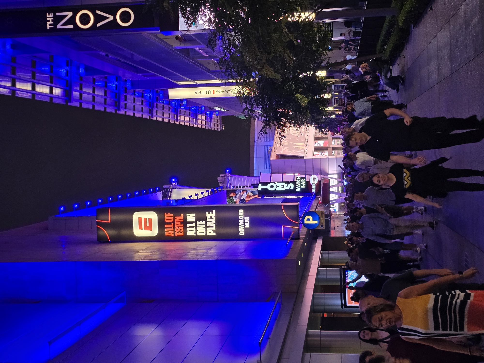 The image depicts an outdoor urban scene at night, likely within the LA Live entertainment district in Los Angeles, California, United States of America. The foreground shows a crowd of people walking on a paved walkway. In the midground and background, large buildings are illuminated with bright blue and purple lights.
Prominent signage is visible, including a large black banner with a red ESPN logo and text that reads "ALL OF ESPN. ALL IN ONE PLACE. DOWNLOAD NOW." Another vertical sign on the right side of the frame displays "THE NOVO" in illuminated white lettering. Below "THE NOVO," a smaller sign reads "ULTRA." A sign for "TOMS WATCH BAR" is also partially visible.
The atmosphere suggests a lively event or gathering is taking place. The people in the crowd are diverse, with various ages and clothing styles, most appearing to be walking in the same direction. Some individuals are engaged in conversation, while others are focused on their path. The lighting from the buildings and signs creates a vibrant and dynamic nocturnal environment. The presence of trees and greenery along the walkway indicates a landscaped urban setting.