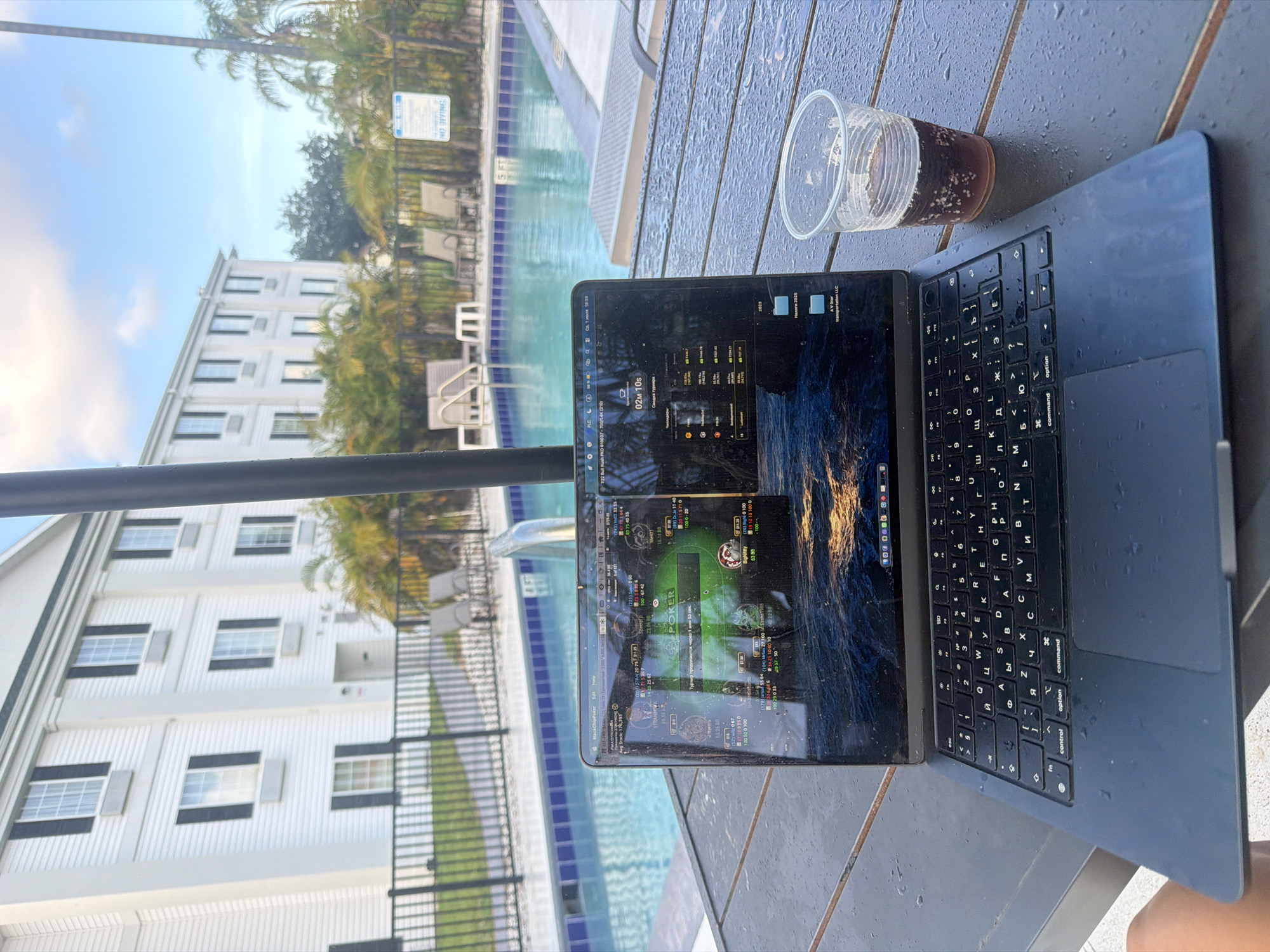 The image shows a laptop open on a wet, dark brown wooden table next to a swimming pool. The laptop screen displays what appears to be a poker game interface with multiple tables visible. Behind the table, a swimming pool with blue water and lounge chairs can be seen. In the background, a white hotel building with multiple windows and balconies is present, along with palm trees. A "NO DIVING" sign is visible near the pool fence. The environment suggests a resort or hotel setting, likely in a warm climate. The time appears to be late afternoon or early evening, given the lighting. There is a plastic cup with a dark beverage and ice cubes next to the laptop. The overall scene depicts someone working or playing poker outdoors by a pool.