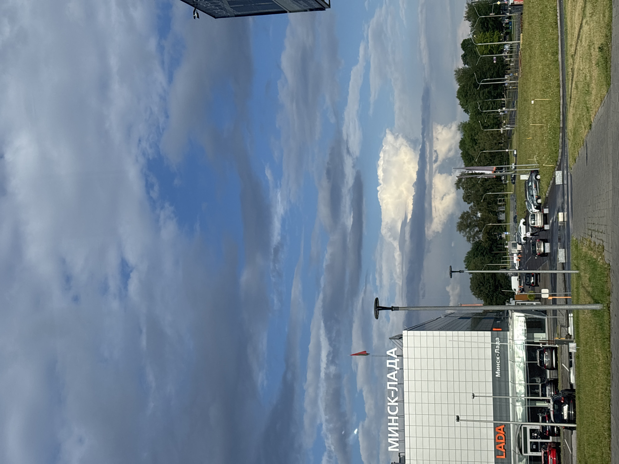 This is a vertical, eye-level, outdoor shot of a car dealership and its surroundings under a dynamic sky. The dealership, identified by "МИНСК-ЛАДА" signage in large white letters on a white, modern building, is prominent on the left. A red and green flag flies from a pole on the roof of the dealership. Below the large signage, smaller "LADA" lettering is visible in orange on the lower portion of the building, with "Минск-Лада" also appearing in white. The building has a glass-fronted showroom area. Parked cars, including a red Lada and several black cars, are visible in the foreground and in the dealership's lot.
The environment includes a paved area in front of the dealership, a grassy field, and a line of trees in the background. A large, cumulonimbus cloud formation dominates the sky, with streaks of dark clouds interspersed with patches of blue sky and brighter, lighter clouds. Streetlights and traffic signals are visible throughout the scene, indicating a road or intersection in the distance. Several cars are driving or parked on a road to the right of the dealership. A person can be seen near the parked cars in front of the dealership, and other figures are present further down the road. The overall scene suggests an overcast but potentially bright day in a suburban or semi-urban area.