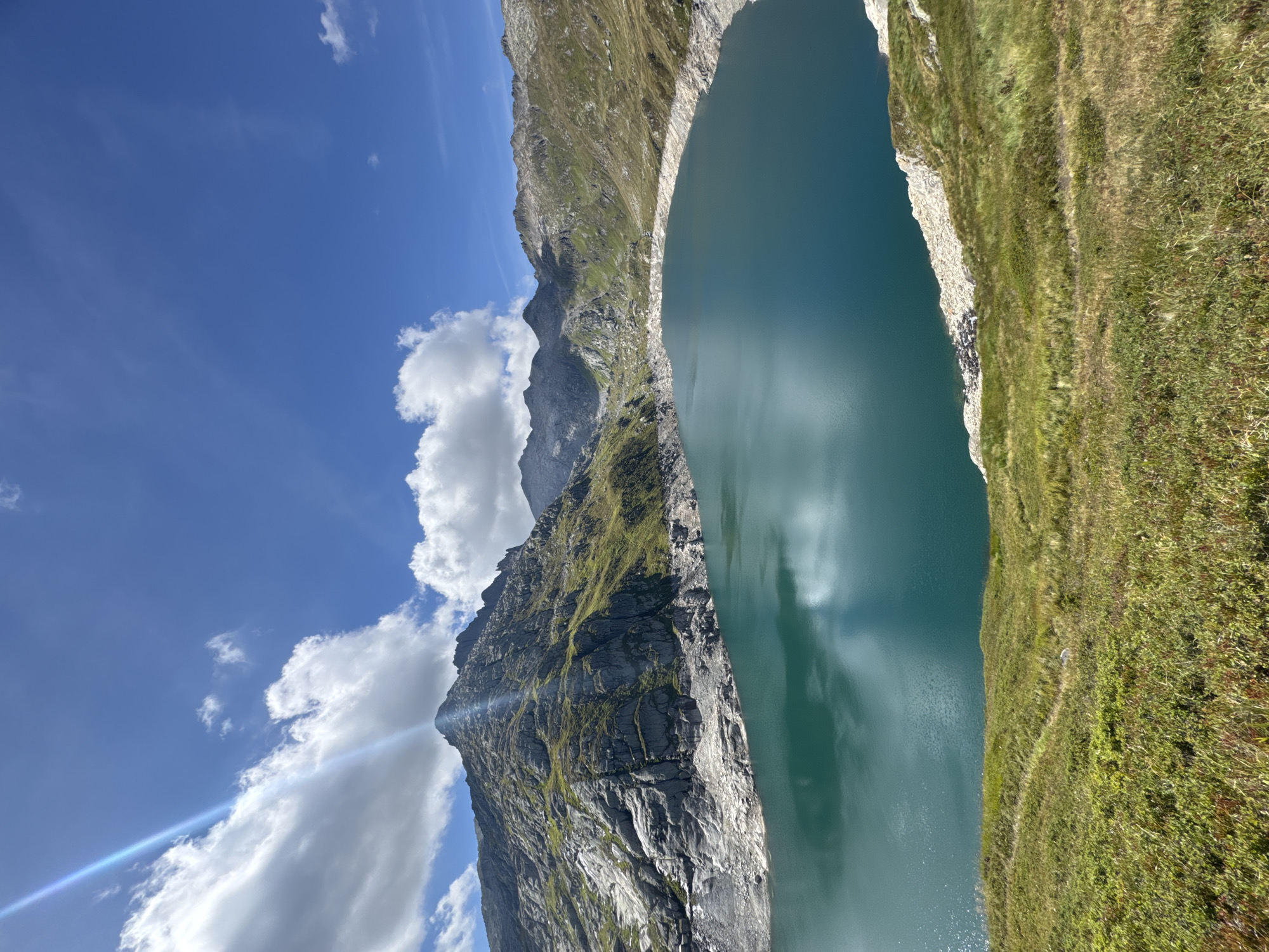 This is a landscape photograph of a reservoir in the mountains near Airolo, Switzerland. The reservoir is a deep turquoise color and is surrounded by rocky, green-sloped mountains. The sky is a bright blue with scattered white clouds. Sunlight streams down from the top left corner of the image, creating a lens flare and casting shadows on the mountains. The foreground is covered in green grass with a winding path visible.