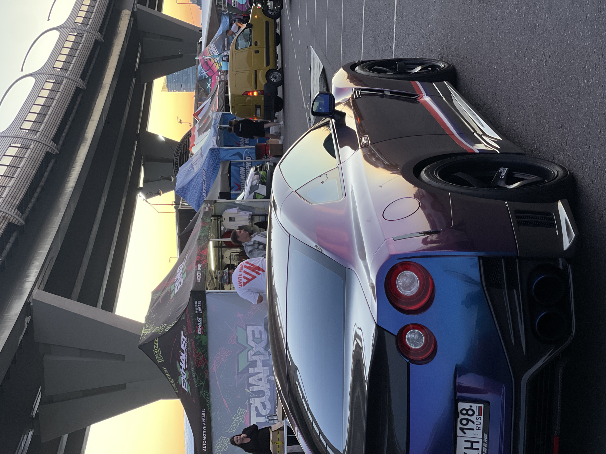 In an outdoor urban environment, a metallic purple and blue Nissan GT-R is parked on asphalt, facing away from the camera and slightly to the right. The car's rear features circular red taillights, dual exhaust pipes, and a license plate that reads "KH 198 RUS". In the background, several tents are set up, displaying logos such as "EXHAUST" and "AUTOMOTIVE APPAREL". A yellow van is parked behind one of the tents. A woman in a black jacket and a man wearing a white t-shirt with red lettering are visible near the tents. Overhead, a large concrete overpass with multiple support pillars stretches across the scene. The sky transitions from a bright orange and yellow at the horizon to a soft blue.