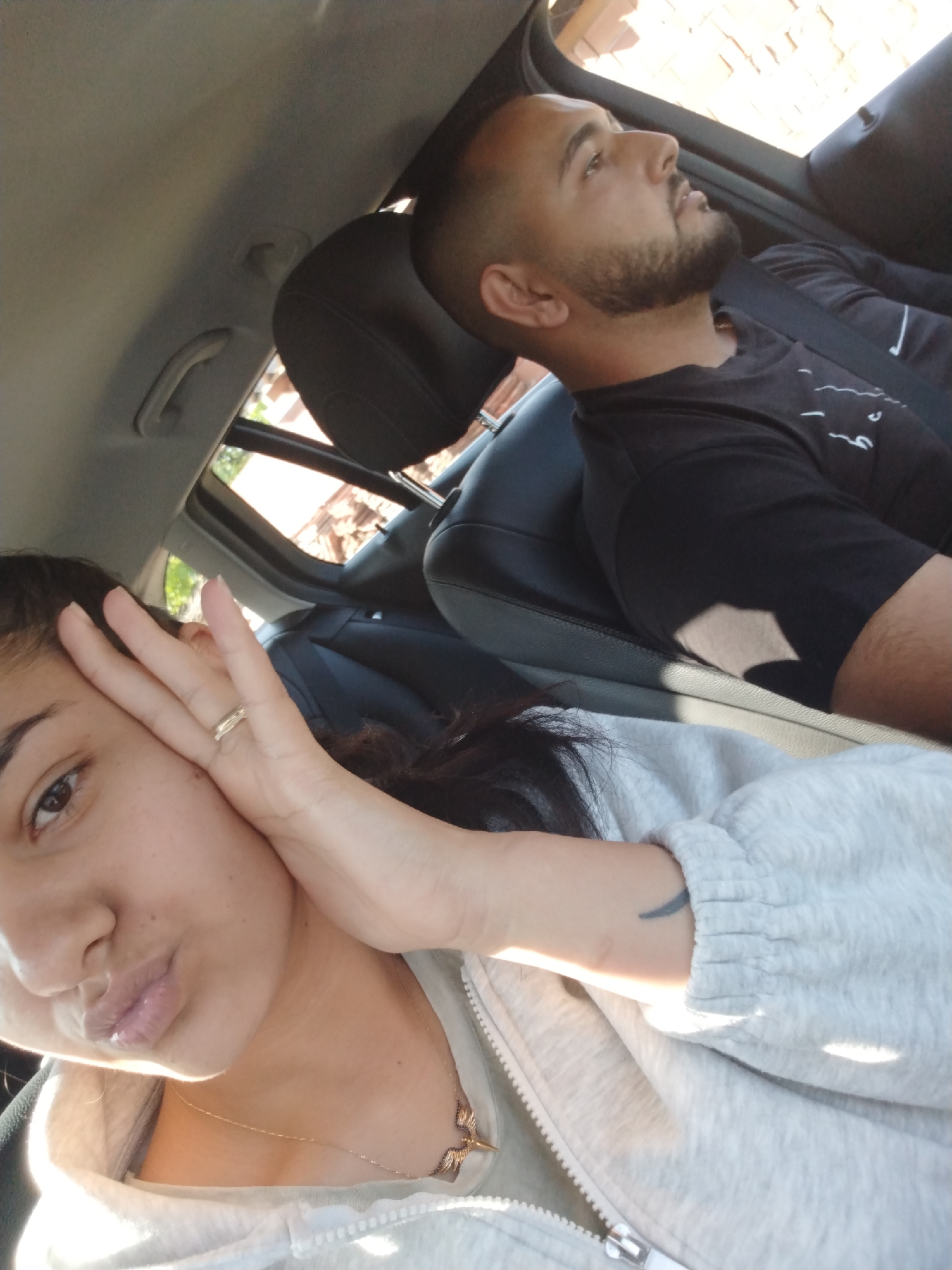 The image captures two individuals inside a car. A woman in the foreground, wearing a gray hooded sweatshirt and a gold necklace, has dark hair. She has her hand near her face, posing. A tattoo is visible on her left wrist. A man is seated in the passenger seat. He has a short haircut and a beard, and wears a black t-shirt. He is looking out the window. The background shows the interior of the car, including the seats, headrest, and part of the car's frame. Sunlight filters through the windows. The location is Unknown, Unknown.