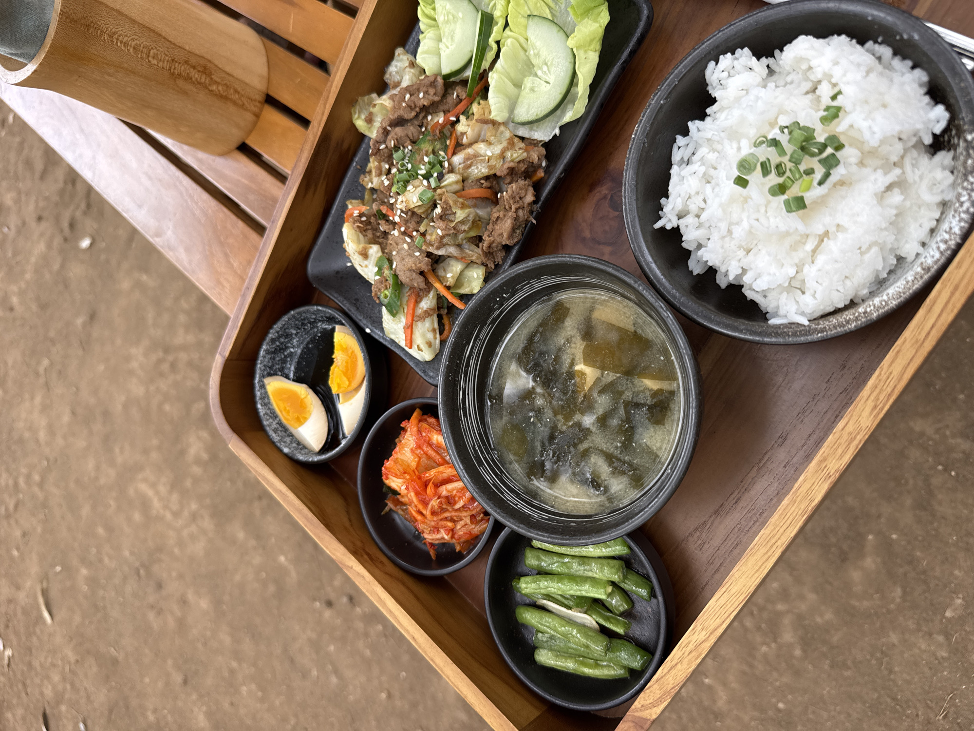 A wooden tray holds a meal consisting of rice, soup, kimchi, green beans, a meat and vegetable stir-fry, and two halves of a hard-boiled egg. The tray is placed on a wooden table. In the background, a tall, cylindrical wooden object is visible. The overall setting suggests an outdoor dining area, possibly a restaurant or cafe, given the rustic wooden elements. The ground visible outside the tray appears to be dirt or sand, consistent with an outdoor environment. The location is Kuta Selatan, Indonesia.