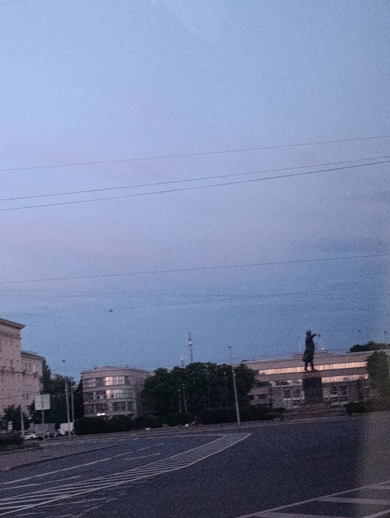 The image depicts a cityscape at dusk or dawn, likely in Saint Petersburg, Russia, given the architectural style of the buildings and the presence of overhead power lines. In the background, a prominent statue of a person stands atop a building. The statue is dark and silhouetted against the lighter sky. The building below the statue has a modern, possibly illuminated facade. To the left, another building with multiple windows is visible. A street with lane markings occupies the foreground, suggesting an urban environment. Trees line the background, interspersed between the buildings, indicating a park or a well-landscaped area. The sky is a gradient of pale blue and hints of pink, typical of twilight. The overall atmosphere is calm and urban.