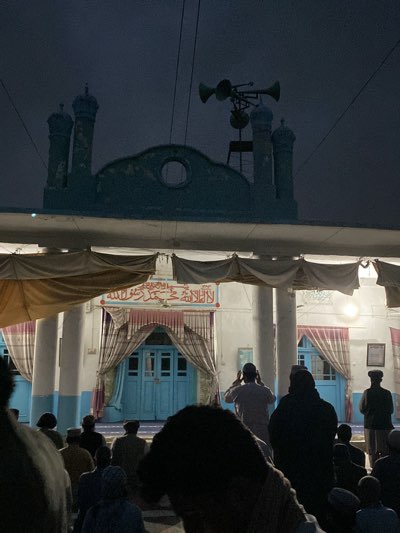 A group of people are gathered in front of a building with an Islamic design, likely a mosque, in Kandahar, Afghanistan. The building is painted white and blue, and has minarets and a dome-like structure on top. A loudspeaker system is mounted on a frame above the mosque. The people are standing and appear to be in prayer or engaged in some religious activity. The scene is dimly lit and seems to be taking place in the evening or night. The men are wearing traditional Afghan attire.