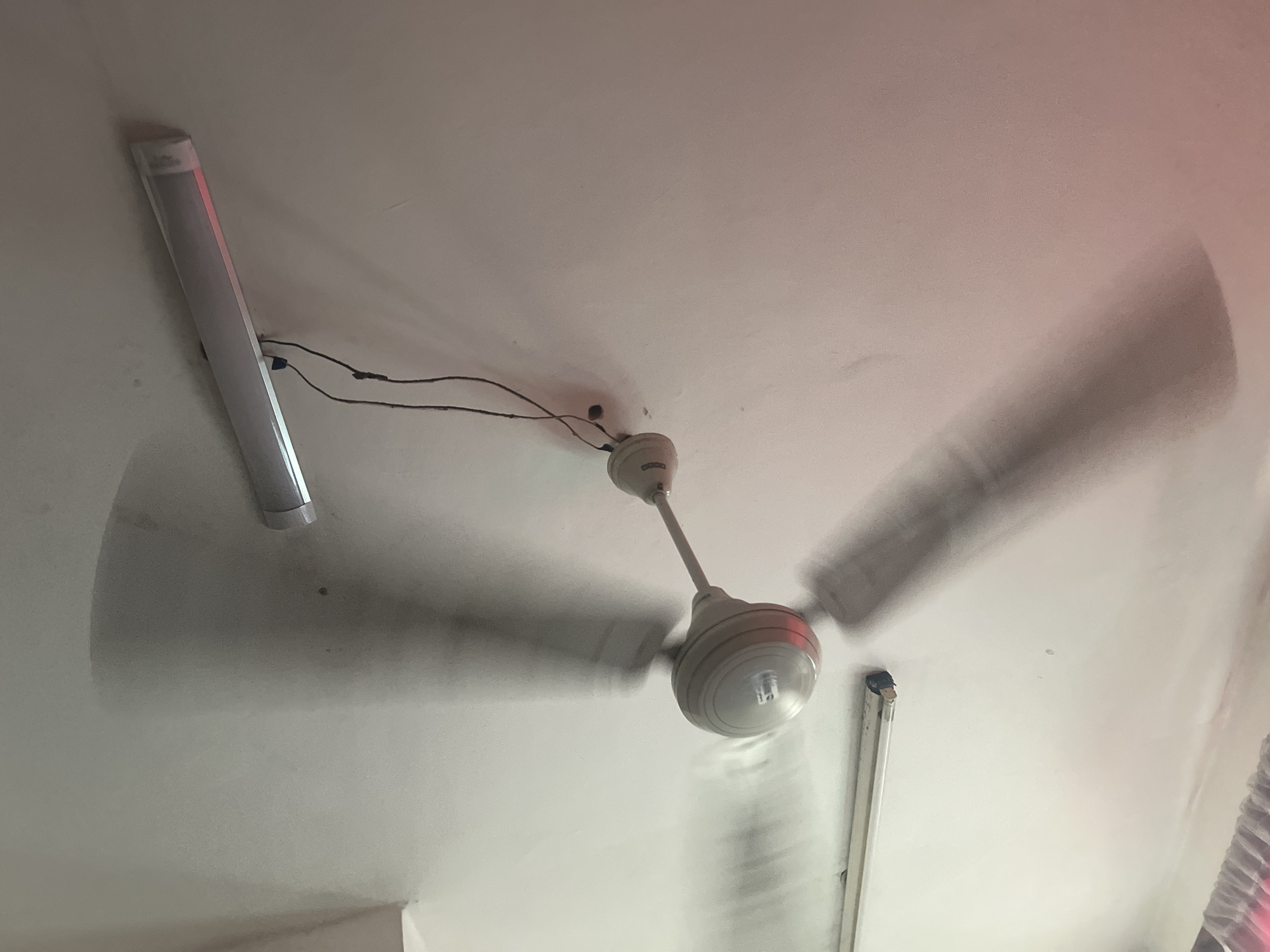The image shows a white ceiling with a rotating ceiling fan in the center. Two fluorescent lights are mounted on the ceiling, one on the left and another on the lower right. Electrical wires are visible connecting the fan to the power source. The blades of the fan appear blurred due to their motion. The environment suggests an indoor space, likely a room in a building. There is no other visual information that indicates the location is in Dar es-Salaam, Tanzania.