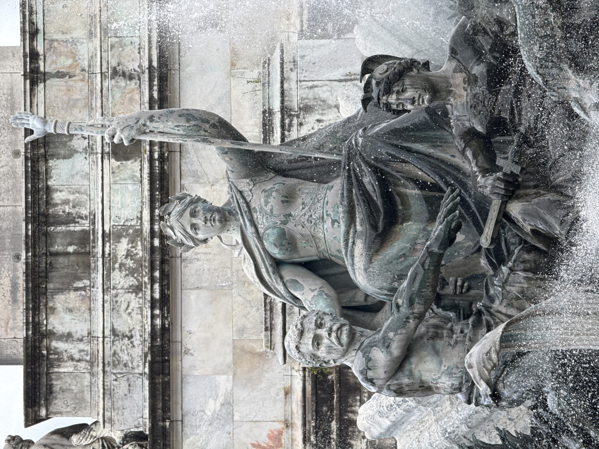 The image shows a close-up of a bronze fountain sculpture in Bordeaux, France, depicting allegorical figures. A central seated female figure, likely representing a deity or personification, holds a staff aloft with her right hand. To her left, a male figure with a mustache and muscular build is positioned. To the right of the central figure, a smaller female figure with a helmet is seated, holding a sword. The sculptures are weathered, with patches of green patina. Water is actively spraying from the fountain, creating a dynamic effect around the figures. Behind the fountain, a stone architectural structure, possibly a monument base, is visible.