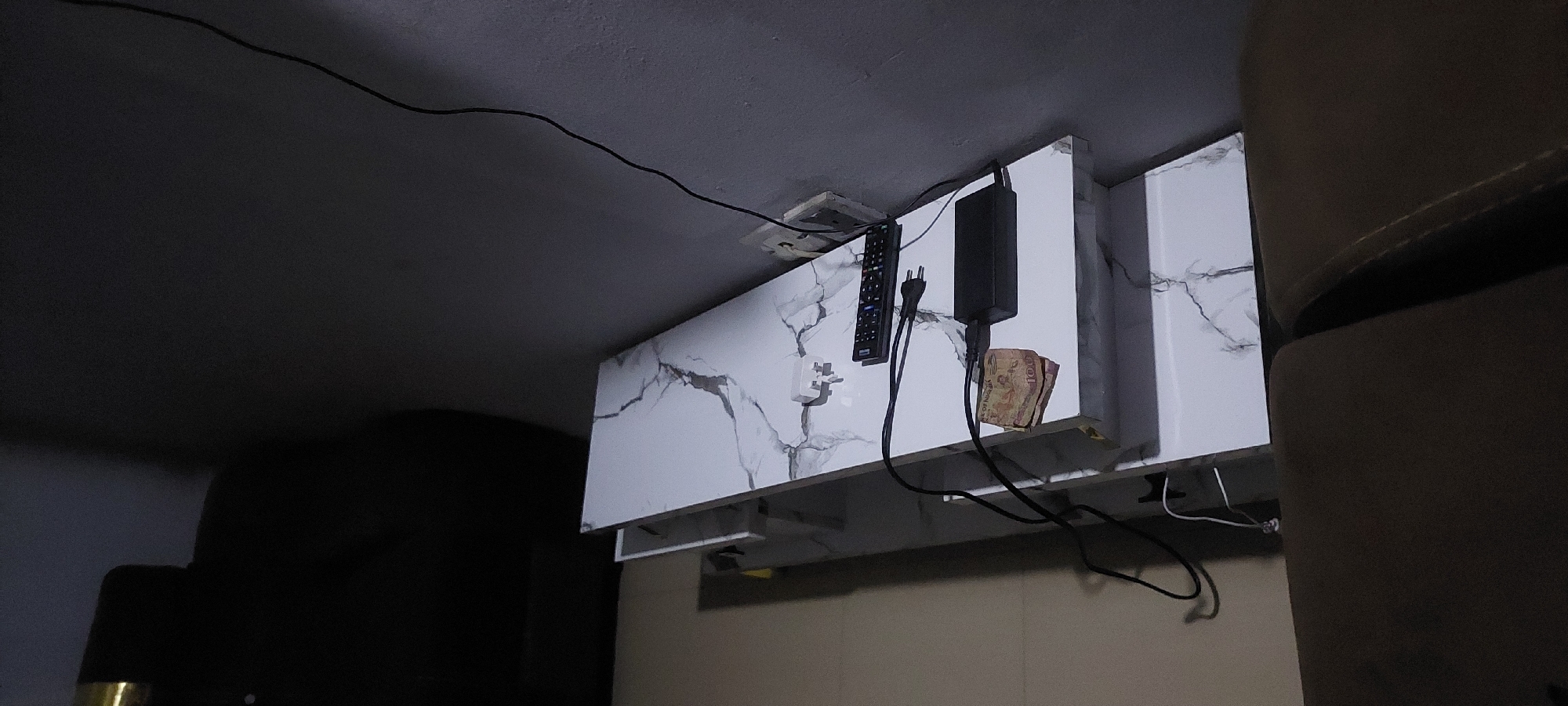 The image shows a modern white marble-patterned television stand in a dimly lit room. On the top shelf of the stand, there is a black remote control, a black power adapter, and a folded 100-unit banknote. There are also two black electrical cords, one plugged into the power adapter and the other leading to the wall outlet. On the wall behind the stand, there are two white electrical outlets. To the left of the stand, a portion of a dark-colored piece of furniture, possibly a sofa or chair, is visible. The floor is covered in light-colored tiles. The overall scene appears to be in a residential setting.